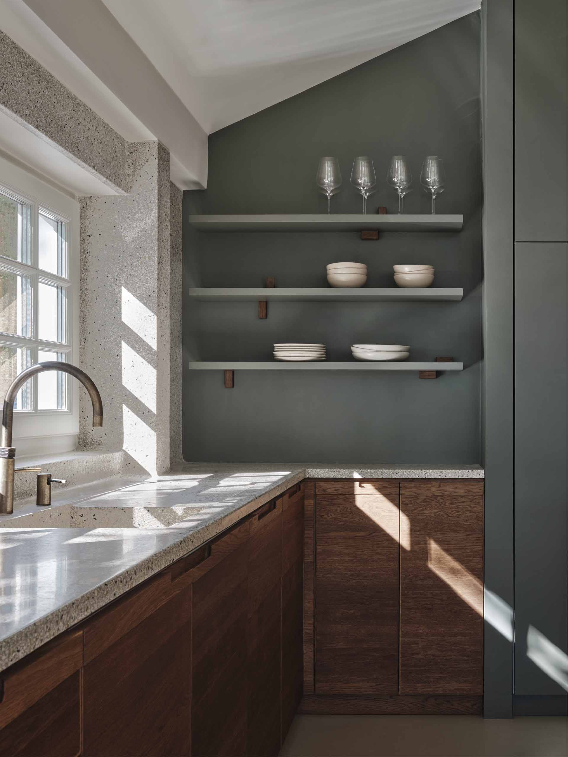 A modern kitchen with dark wood and matte green cabinets, also includes a seamless terrazzo countertop and integrated sink, that then travels up onto the wall.