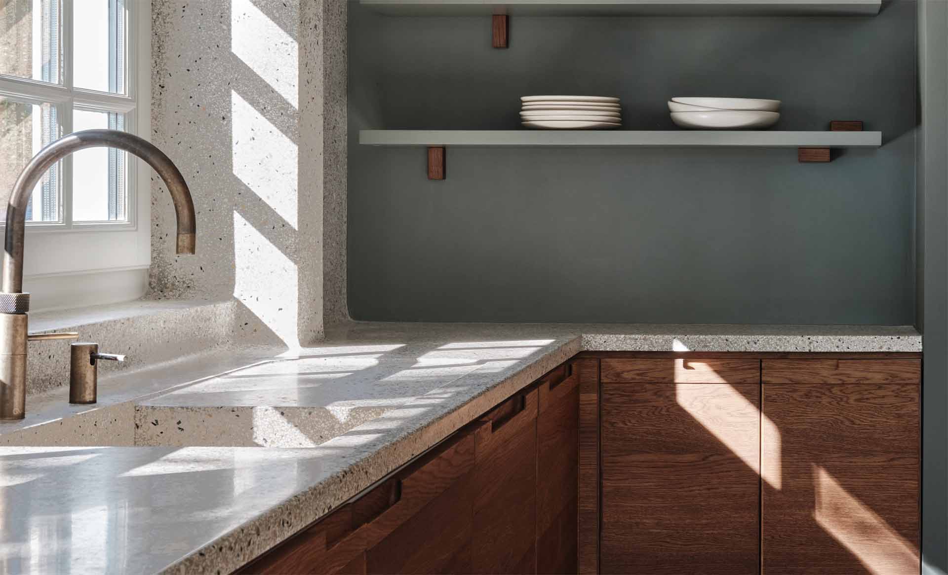 A modern kitchen with dark wood and matte green cabinets, also includes a seamless terrazzo countertop and integrated sink, that then travels up onto the wall.