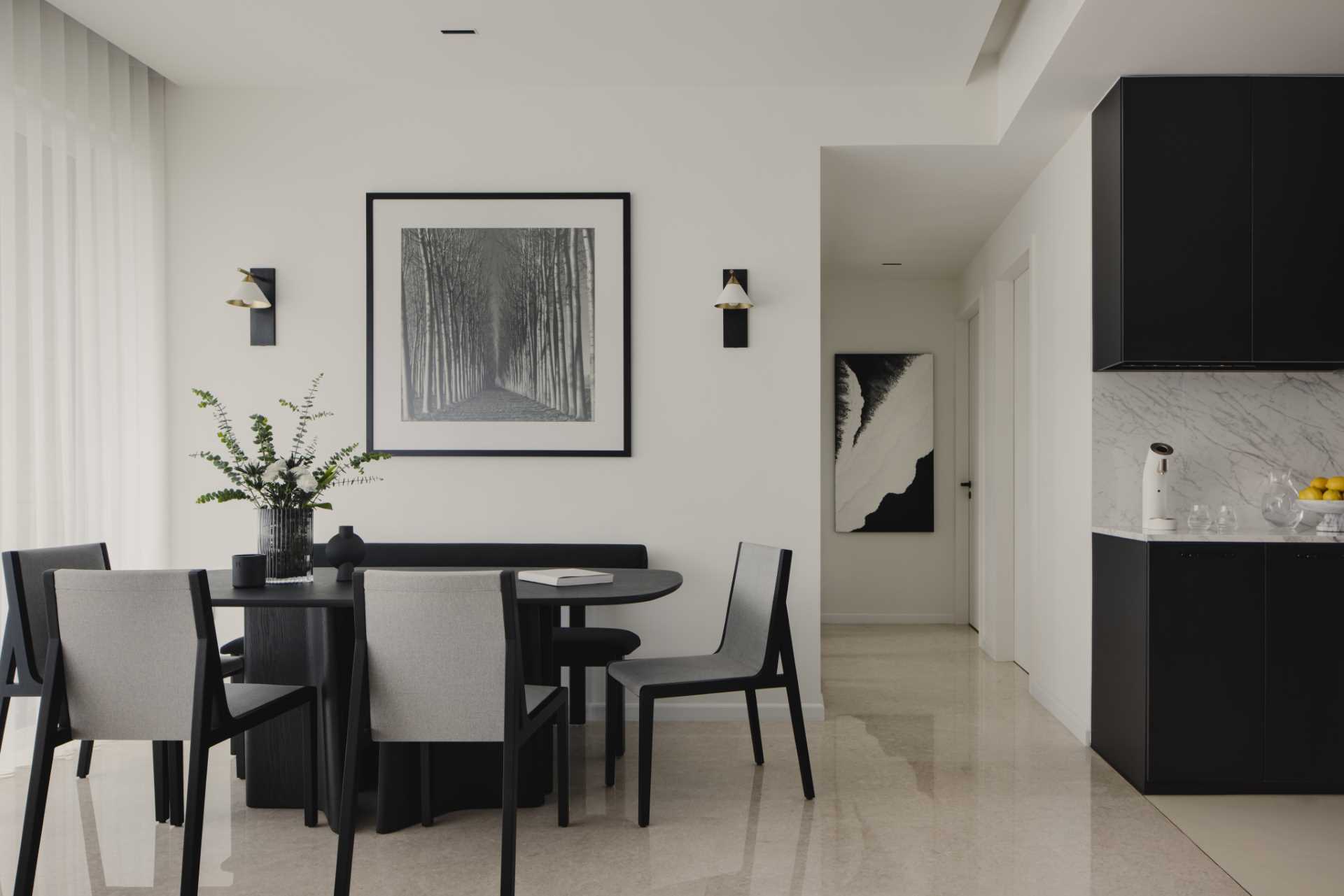 This modern and monochrome dining area is located behind the living room and includes a black dining table with a sculptural base, black and grey dining chairs, a black bench, black and white artwork, and wall lights with a brushed gold accent.