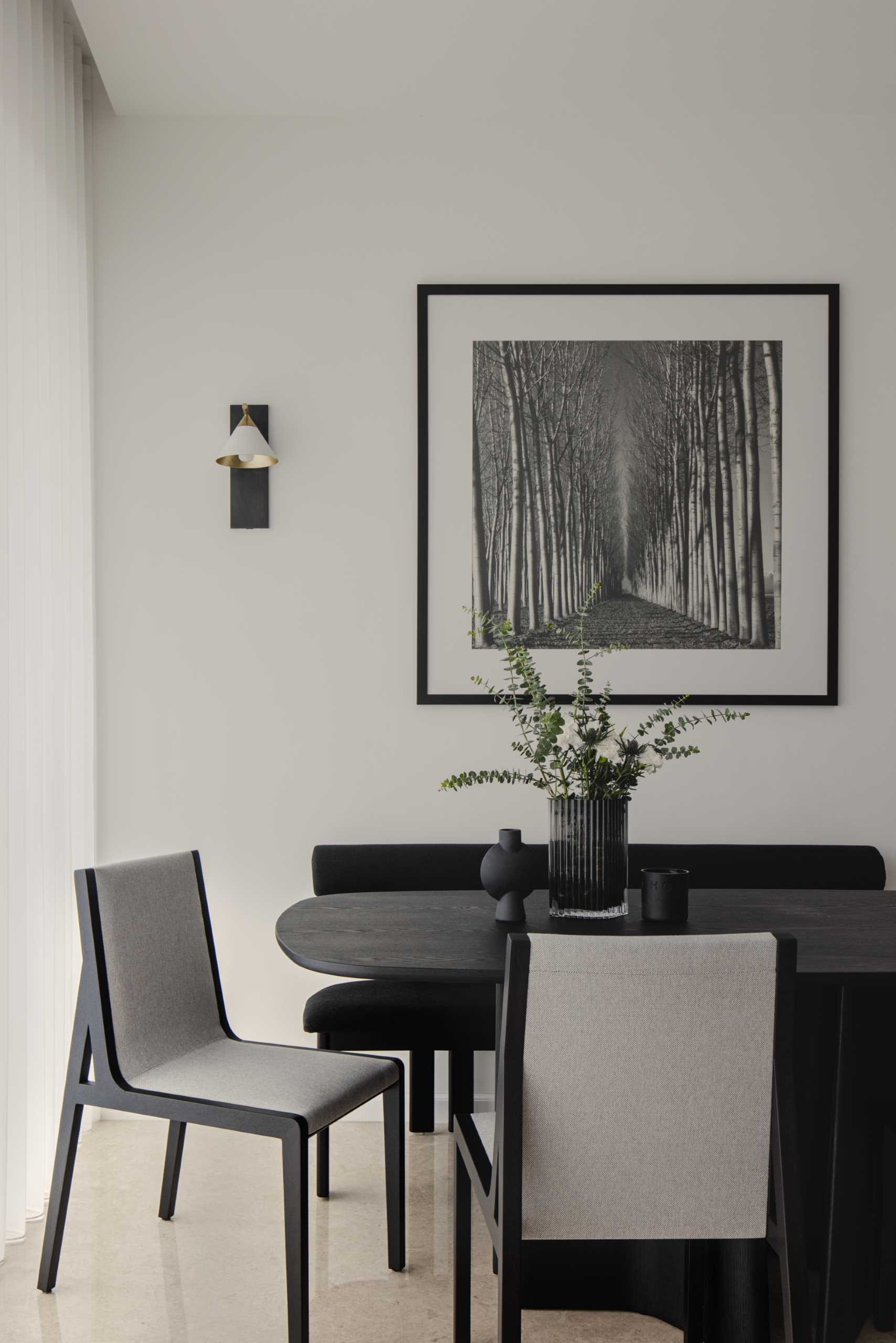 This modern and monochrome dining area is located behind the living room and includes a black dining table with a sculptural base, black and grey dining chairs, a black bench, black and white artwork, and wall lights with a brushed gold accent.