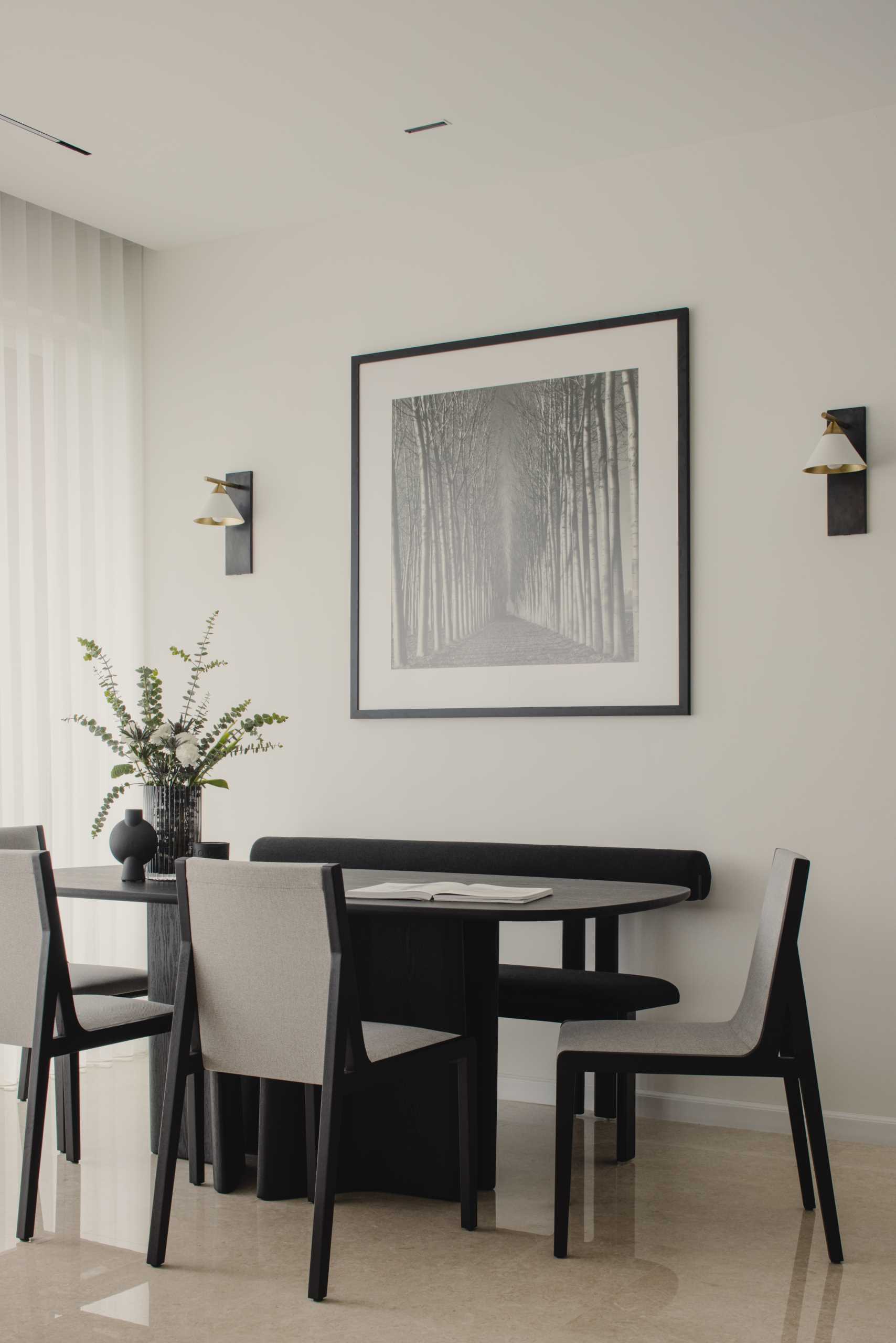 This modern and monochrome dining area is located behind the living room and includes a black dining table with a sculptural base, black and grey dining chairs, a black bench, black and white artwork, and wall lights with a brushed gold accent.