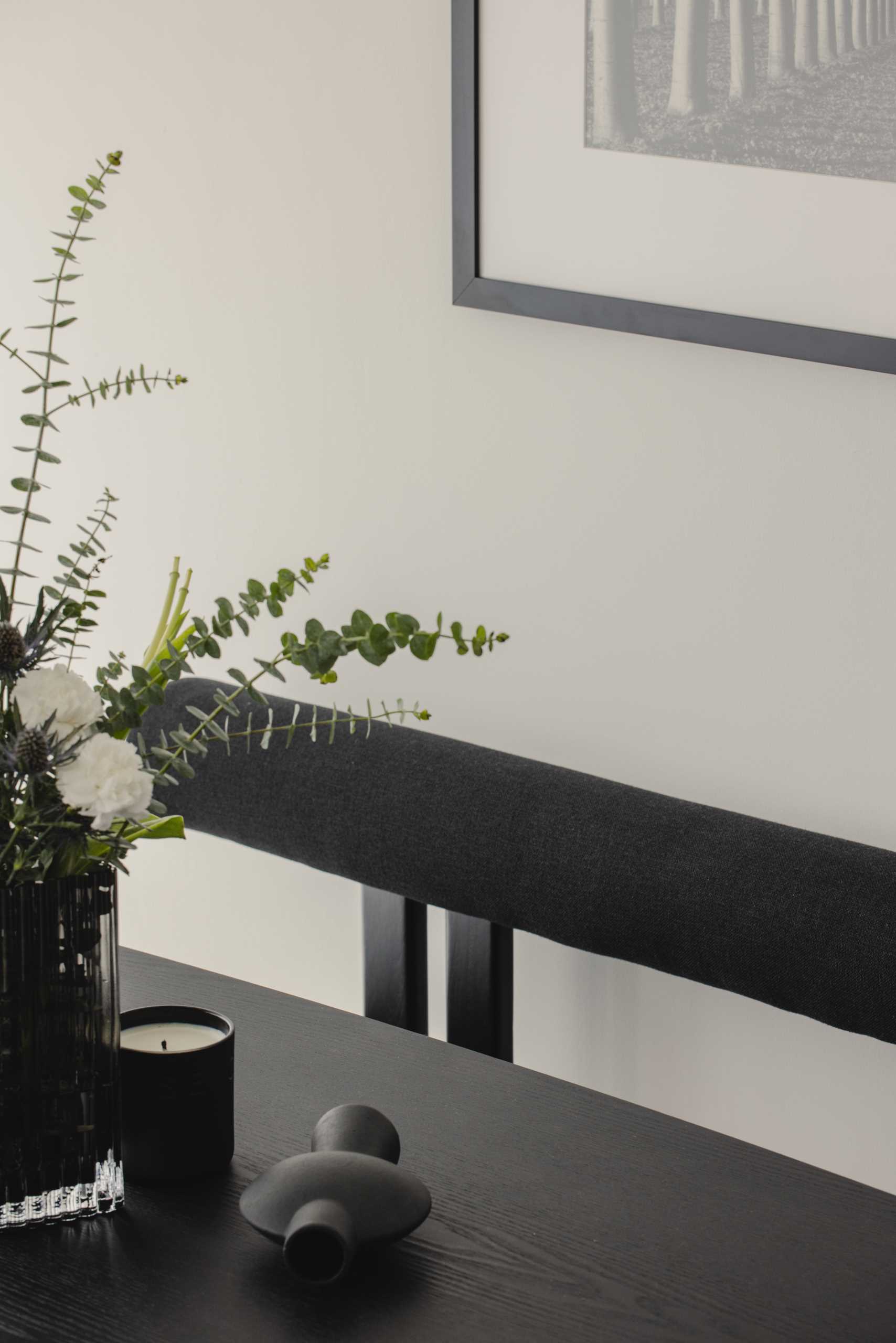 This modern and monochrome dining area is located behind the living room and includes a black dining table with a sculptural base, black and grey dining chairs, a black bench, black and white artwork, and wall lights with a brushed gold accent.