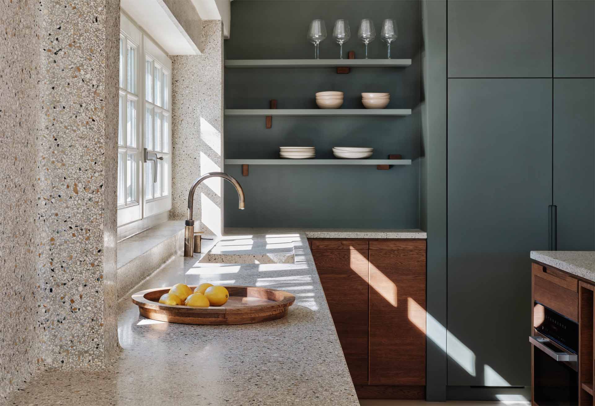 A modern kitchen with dark wood and matte green cabinets, also includes a seamless terrazzo countertop and integrated sink, that then travels up onto the wall.