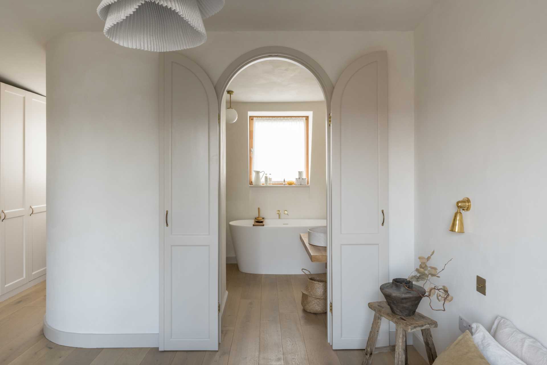 An arched doorway leads from the bedroom to the ensuite bathroom, where there's a freestanding bathtub under the window, a wood vanity, and a shower with a built-in seat.