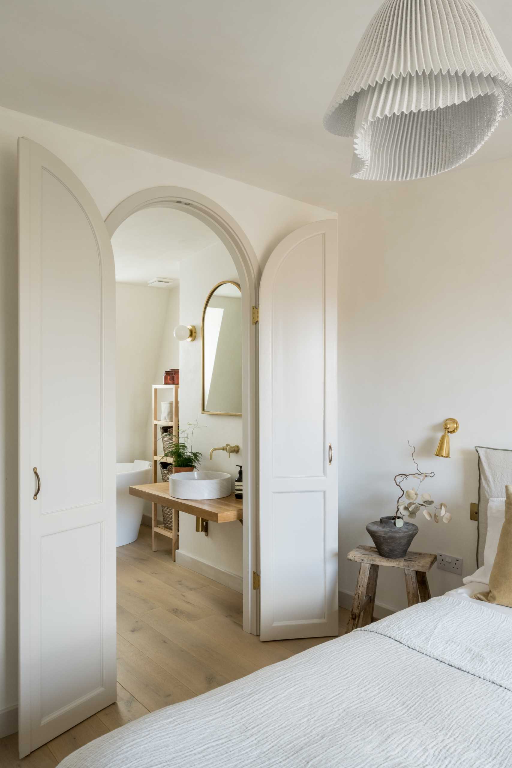 An arched doorway leads from the bedroom to the ensuite bathroom, where there's a freestanding bathtub under the window, a wood vanity, and a shower with a built-in seat.