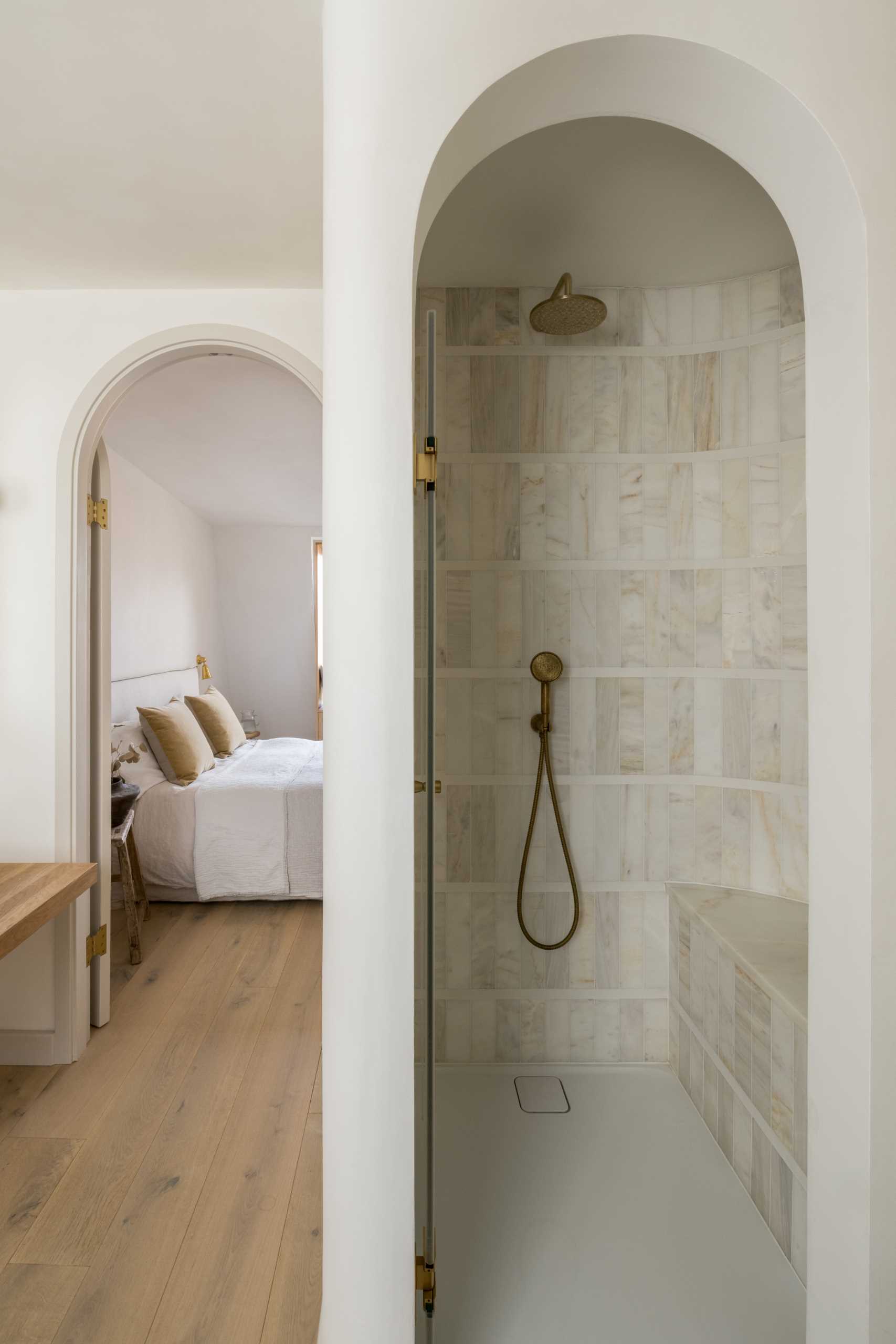 An arched doorway leads from the bedroom to the ensuite bathroom, where there's a freestanding bathtub under the window, a wood vanity, and a shower with a built-in seat.