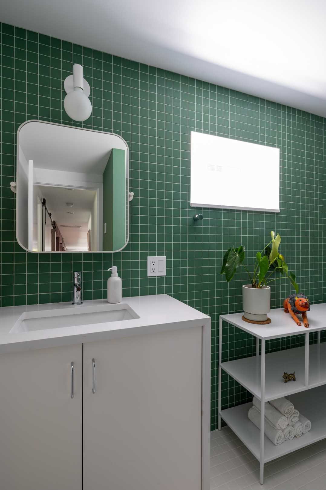 A white and emerald green guest bath, that features small green tiles and a white open shelving unit.