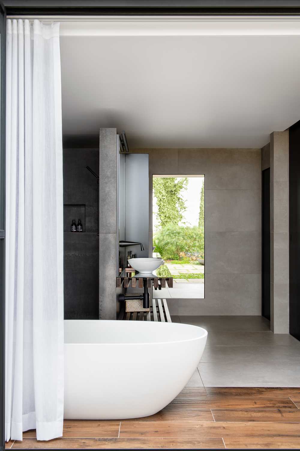 In this modern bathroom, there's a freestanding bathtub, a vanity with open wood shelving, a walk-in shower is positioned behind the wall, and a mirror reflecting the view of the garden.