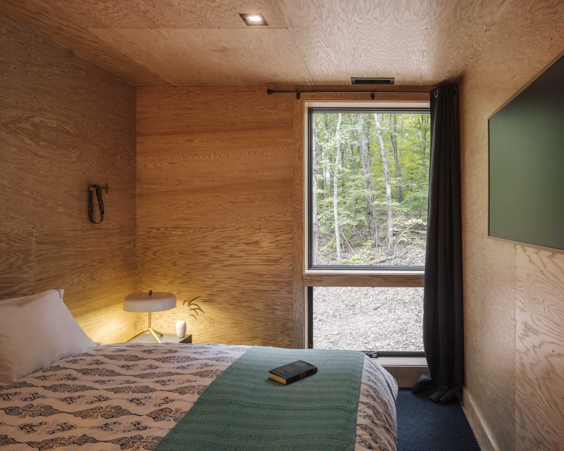 A bedroom lined with wood include a vertical window for natural light and views of the trees.