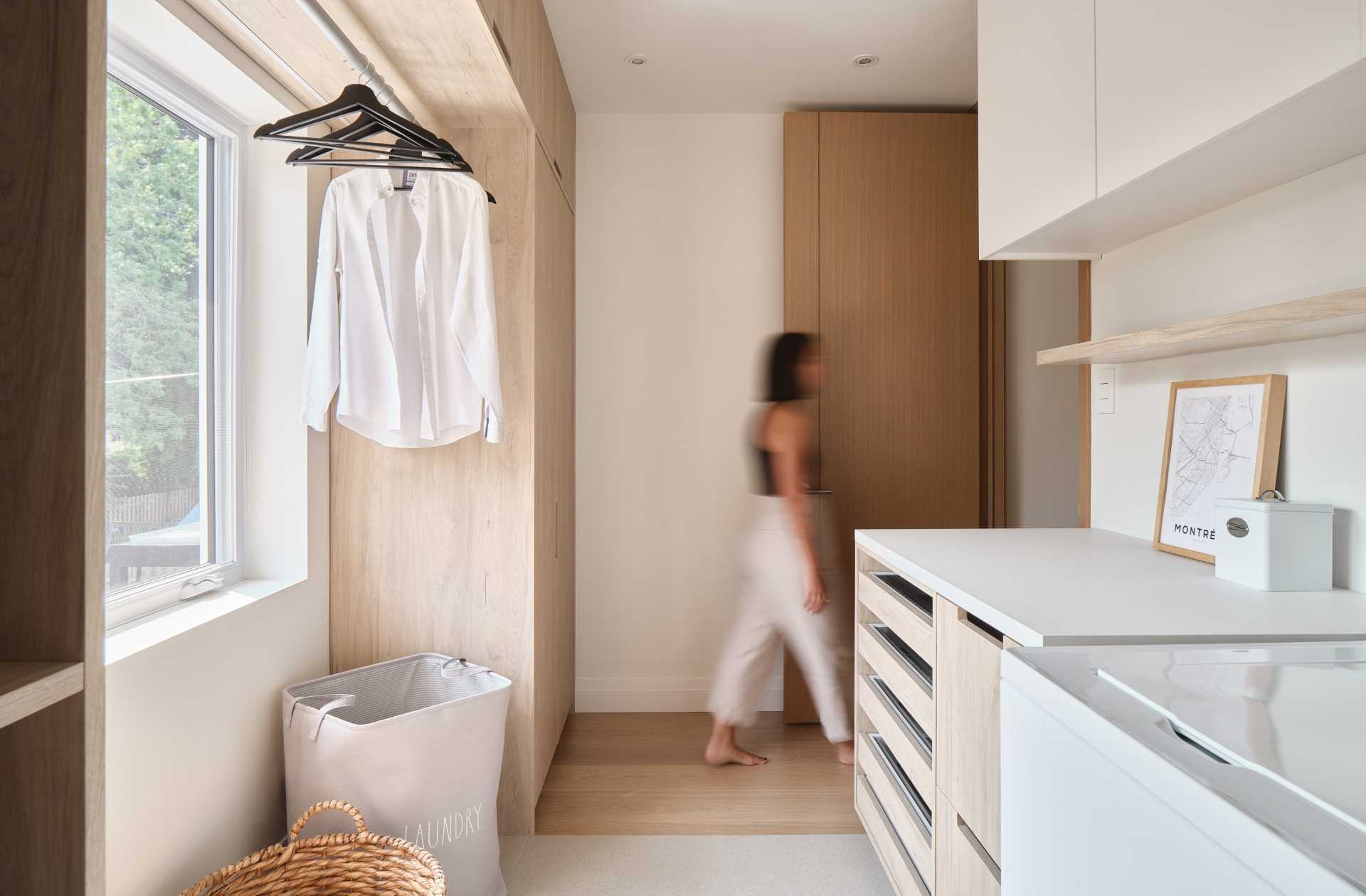 This modern laundry room includes cabinets and shelving that wrap around the window, with a clothing rod located in front of the window to dry clothes.