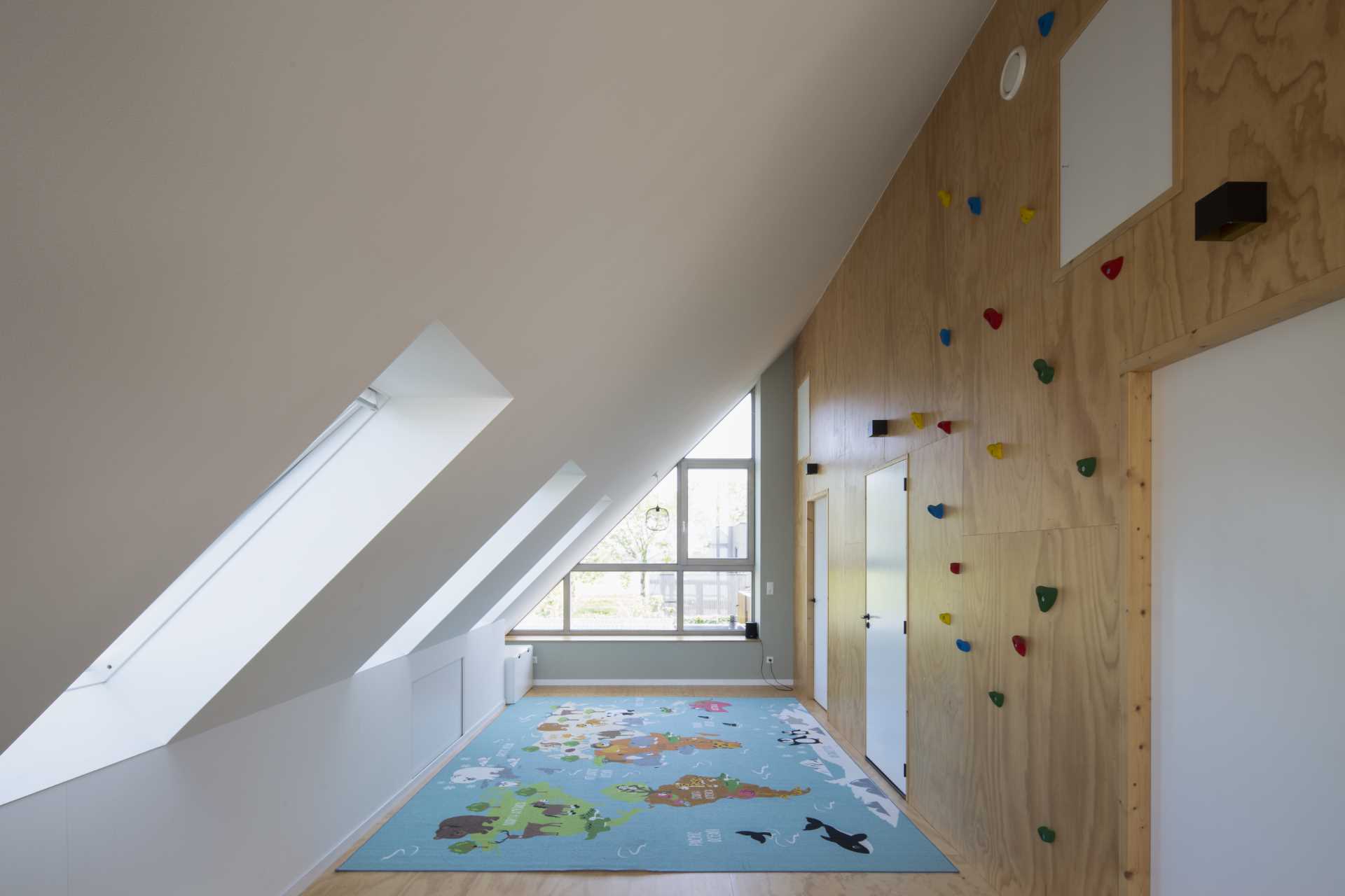 The playroom, with its sloped ceiling, has a plywood wall adorned with rock climbing supports, while a colorful rug covers the floor. The white doors in the plywood wall lead to bedrooms and a bathroom.