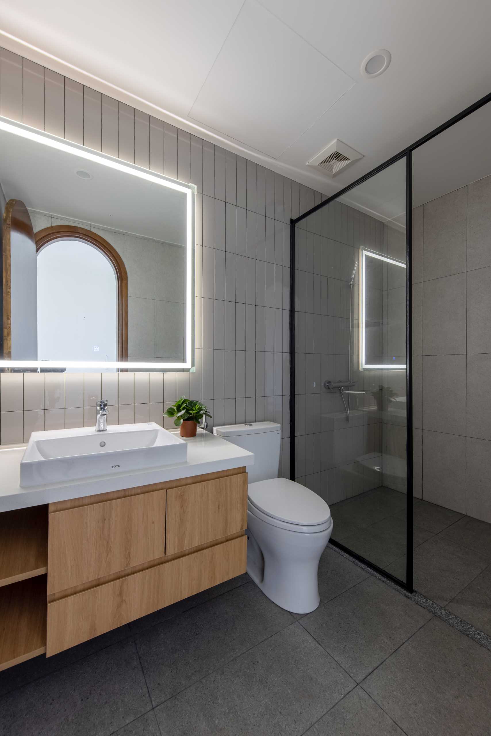 This primary ensuite bathroom includes walls covered in vertical tiles, a black-framed s،wer, and a wood vanity with a white countertop.