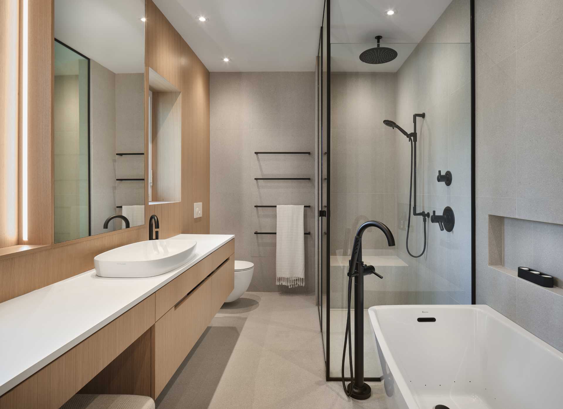 In this modern ensuite bathroom, there's a large vanity, a freestanding bathtub with nearby shelving niche, and a black-framed glass-enclosed shower.