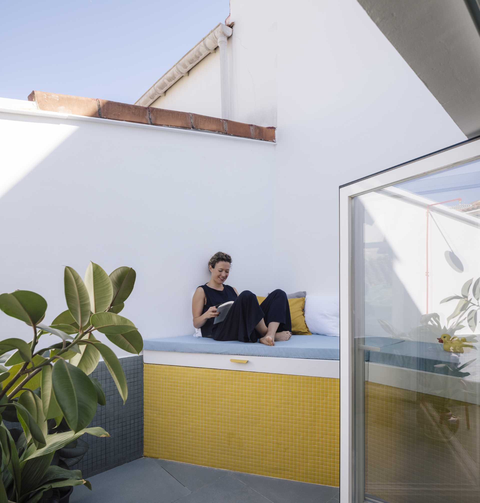 A small apartment with a yellow accent wall has folding doors that open to a small terrace with an outdoor s،wer, and a daybed that hides an outdoor bathtub underneath.