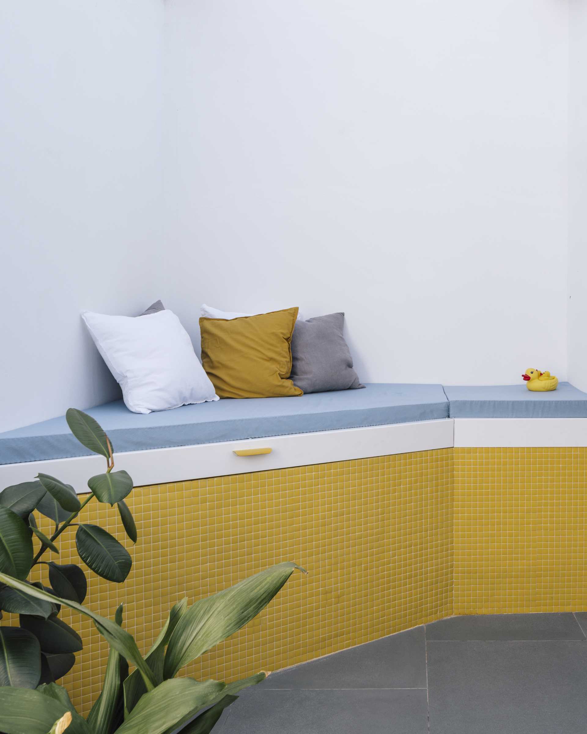 A small apartment with a yellow accent wall has folding doors that open to a small terrace with an outdoor s،wer, and a daybed that hides an outdoor bathtub underneath.