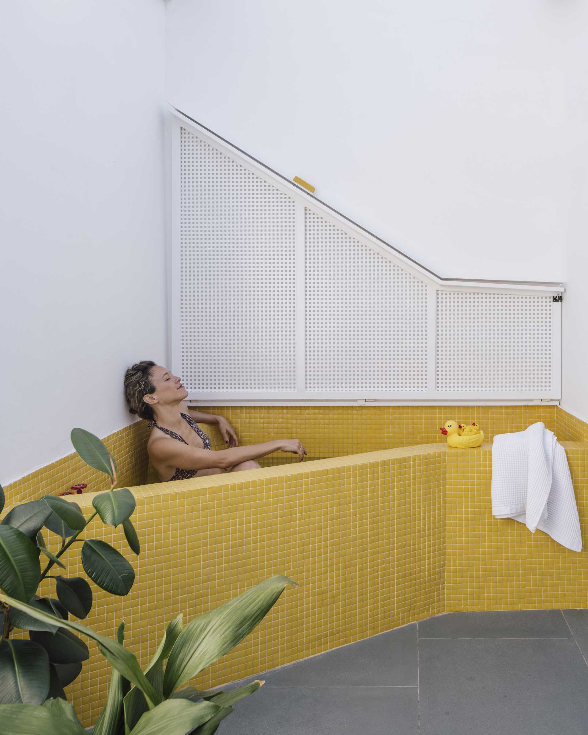 A small apartment with a yellow accent wall has folding doors that open to a small terrace with an outdoor s،wer, and a daybed that hides an outdoor bathtub underneath.