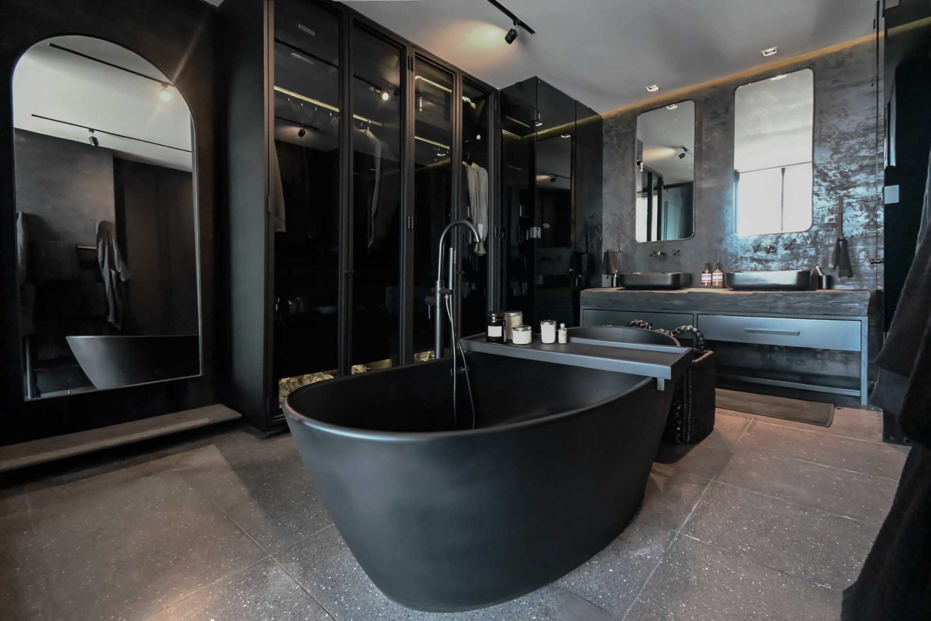 A modern black bathroom includes a freestanding black bathtub in the center of the room, a wall of glass-fronted closets, and a double vanity. The toilet and shower are located behind glass boxes on either side of the vanity.