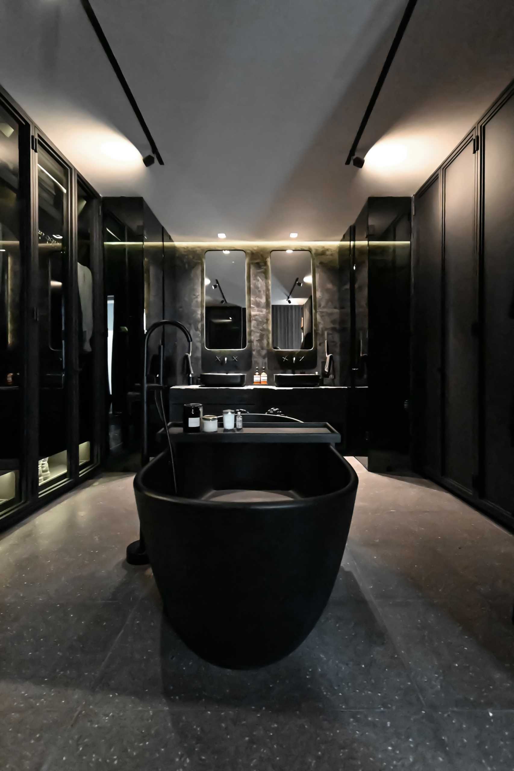 A modern black bathroom includes a freestanding black bathtub in the center of the room, a wall of glass-fronted closets, and a double vanity. The toilet and shower are located behind glass boxes on either side of the vanity.