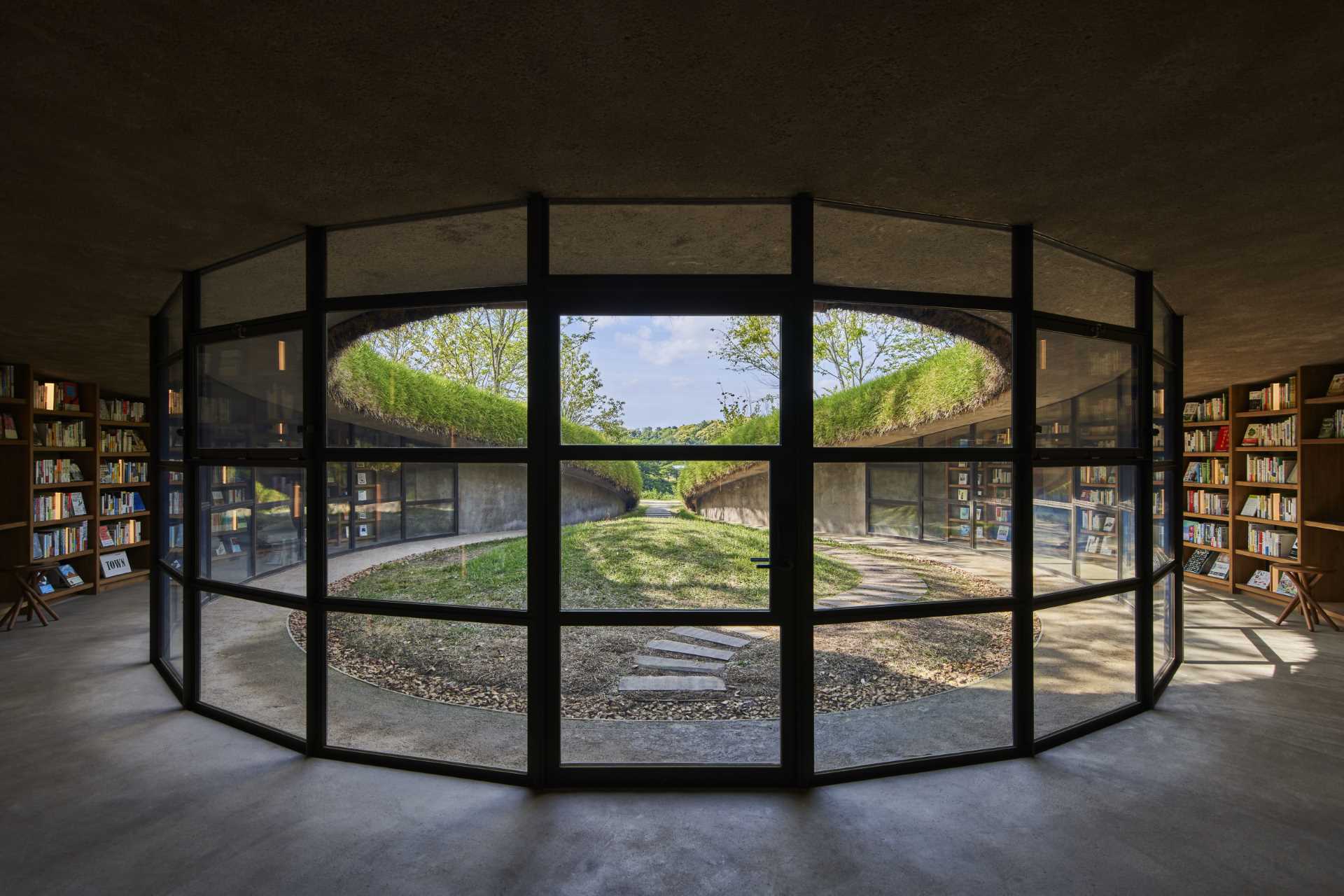 A modern library built into the earth includes a curved glass wall and bookshelves that wrap around the wall.