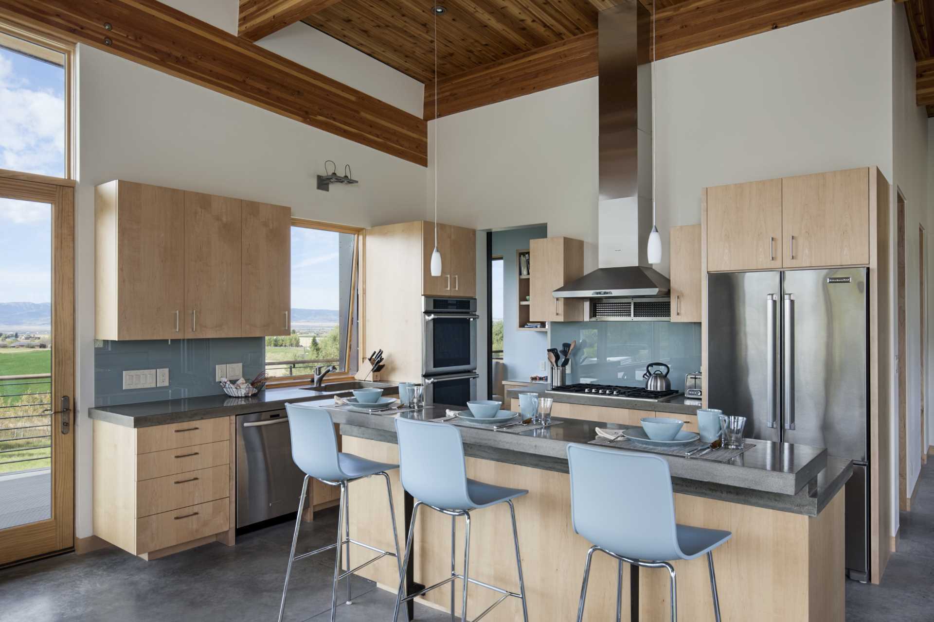 In the kitchen, light wood cabinets have been paired with a colored glass backsplash, concrete countertops, and stainless steel appliances.