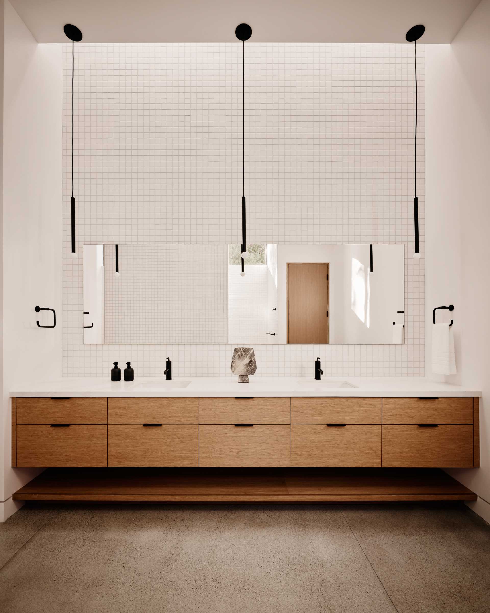 In this modern bathroom, tiles cover the walls, minimalist black pendant lights hang from the ceiling, and a wood vanity spans the wall.