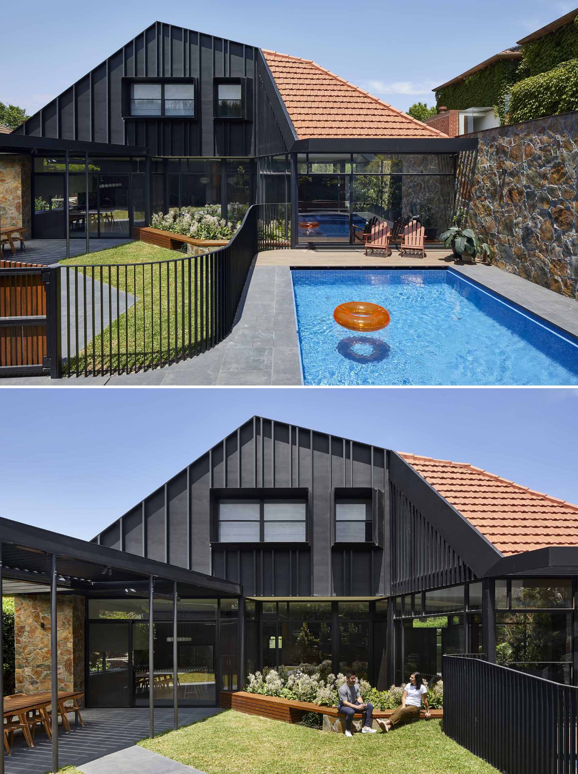 The rear exterior of this modern home features a black facade and a stone wall, while a black fence separates the swimming pool from the yard, where a pergola provides shade for outdoor dining, and a wood bench wraps around a planter.