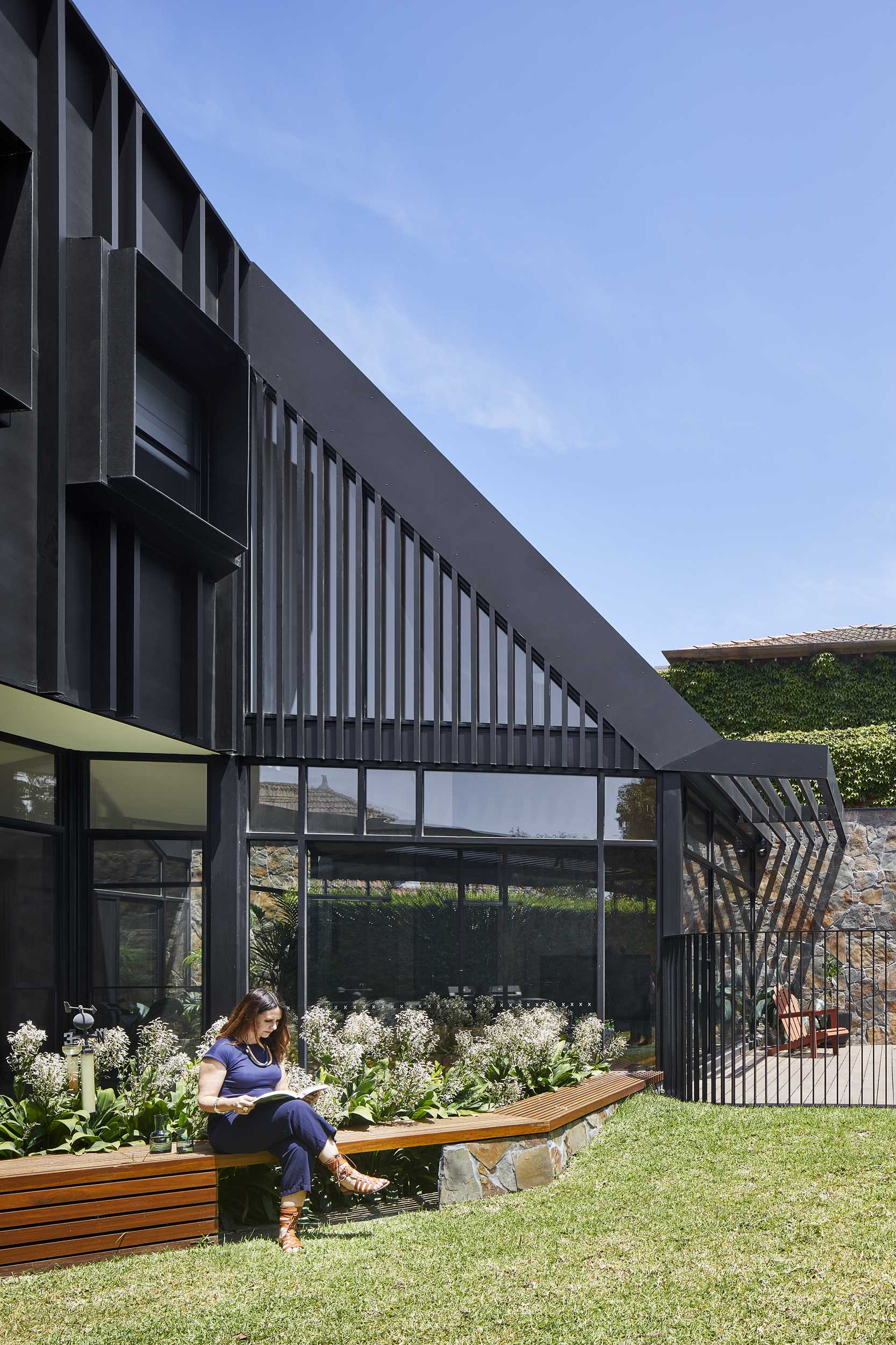 The rear exterior of this modern home features a black facade and a stone wall, while a black fence separates the swimming pool from the yard, where a pergola provides shade for outdoor dining, and a wood bench wraps around a planter.