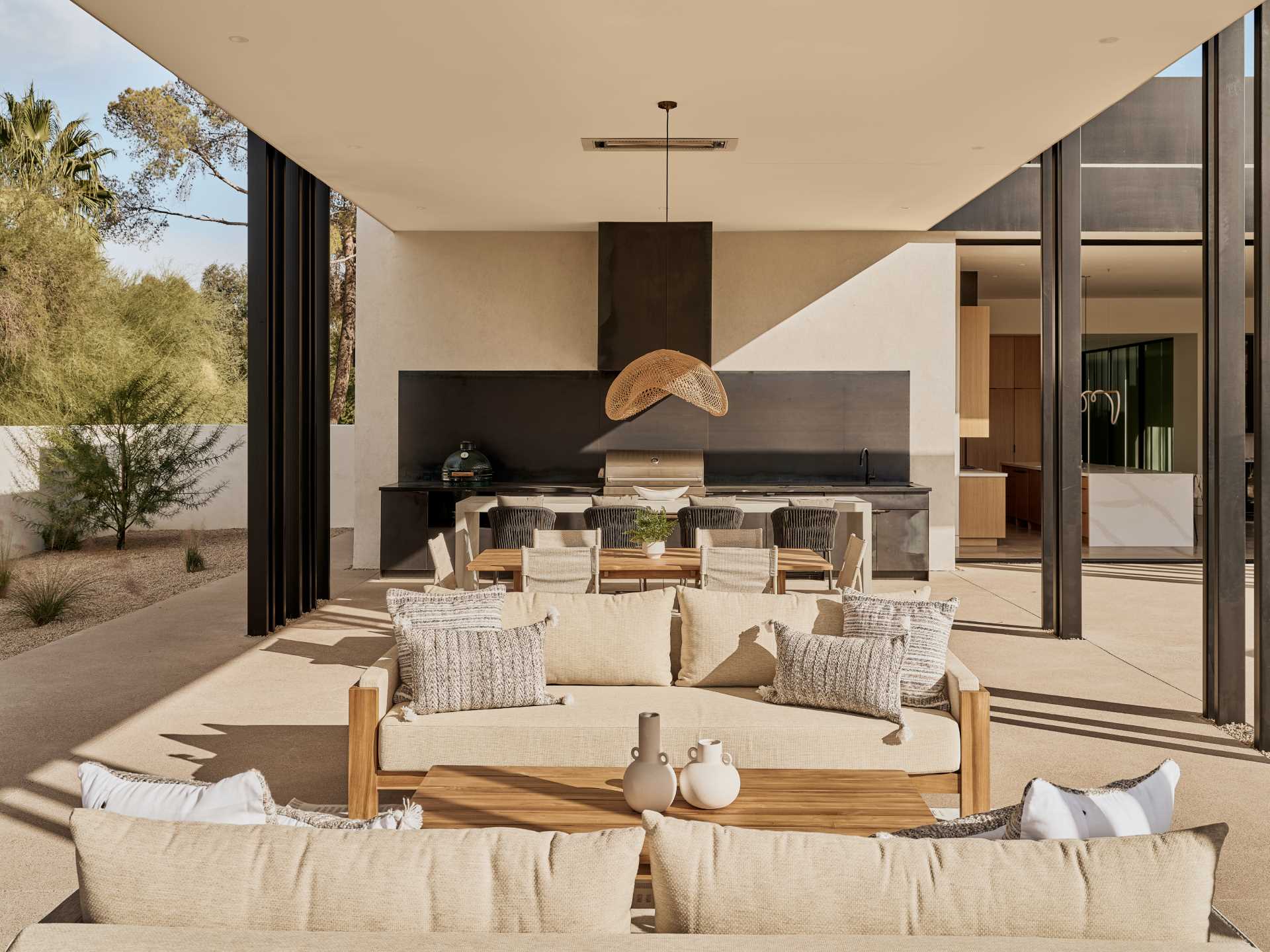 An expansive covered patio supported by thin metal fins and anchored by a steel fireplace features a full kitchen with a poured concrete island, and dining and living areas.