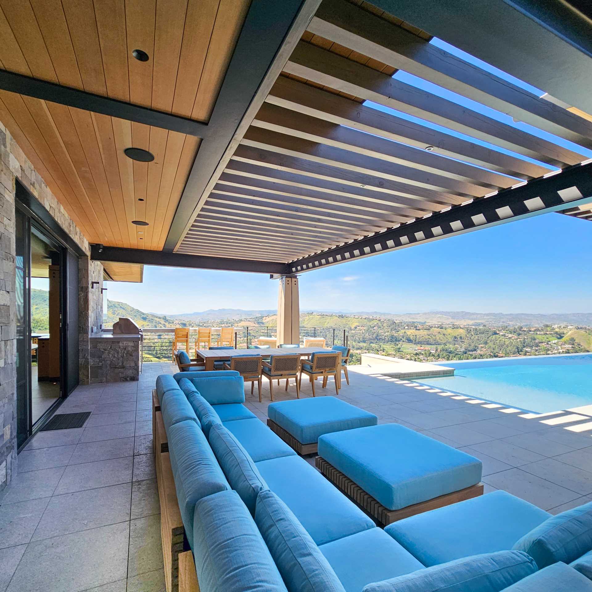 Wood and steel columns are on display by the pool of this contemporary house, while the overhanging roof provides shelter and shade, and a pergola defines an outdoor living space furnished with a couch, dining area, and bbq.