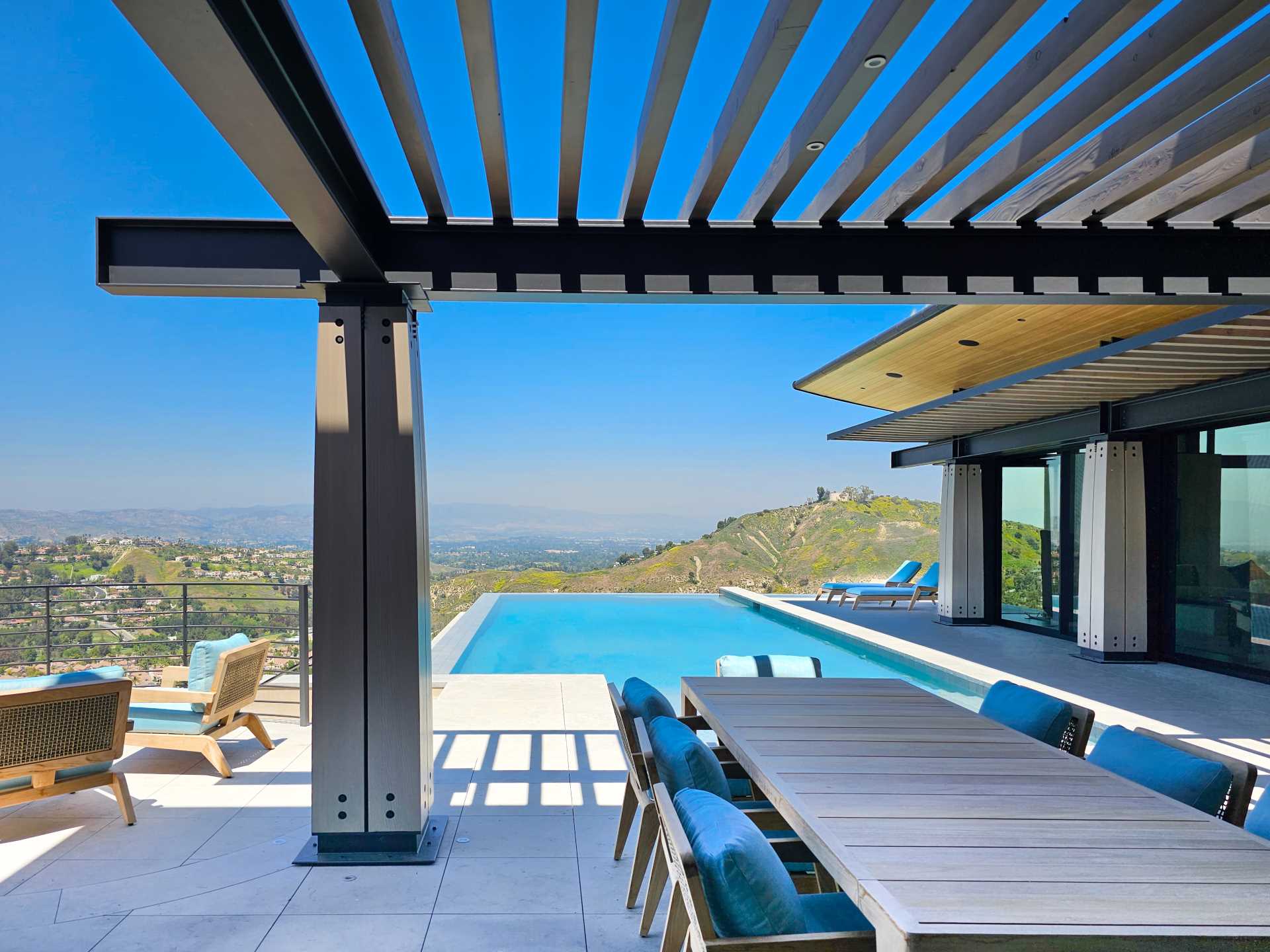 Wood and steel columns are on display by the pool of this contemporary house, while the overhanging roof provides shelter and shade, and a pergola defines an outdoor living space furnished with a couch, dining area, and bbq.