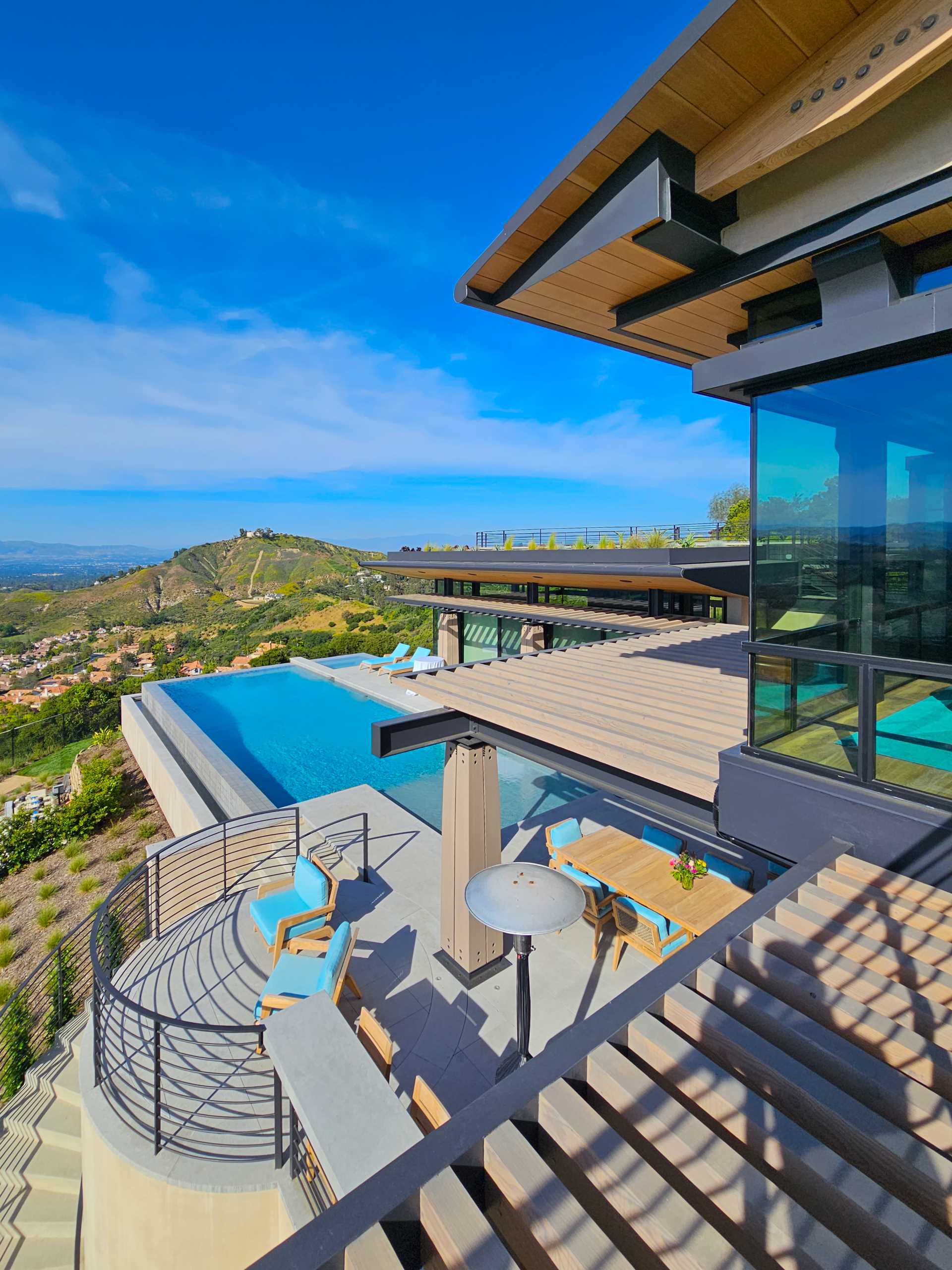 Wood and steel columns are on display by the pool of this contemporary house, while the overhanging roof provides shelter and shade, and a pergola defines an outdoor living space furnished with a couch, dining area, and bbq.