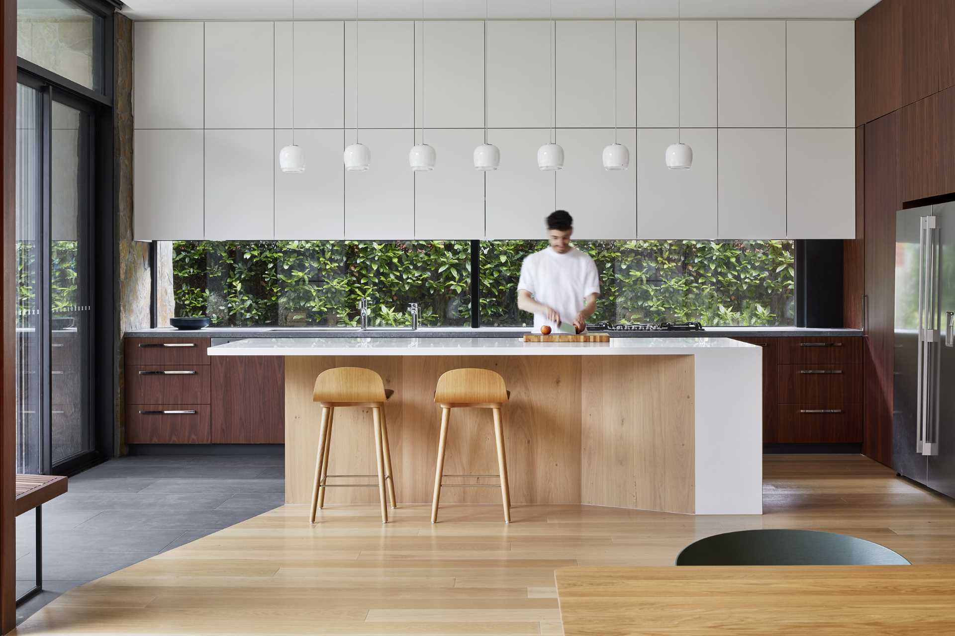 In this modern kitchen, dark wood cabinetry has been paired with minimalist white cabinets, while the island has room for a pair of stools and seven pendant lights to ensure the island is well-lit. The kitchen also includes a gl، backsplash, allowing the plants outside to be seen.
