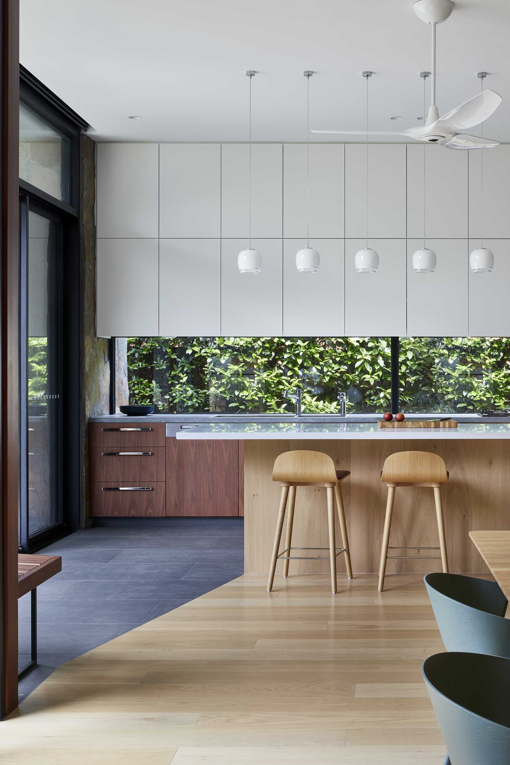 In this modern kitchen, dark wood cabinetry has been paired with minimalist white cabinets, while the island has room for a pair of stools and seven pendant lights to ensure the island is well-lit. The kitchen also includes a glass backsplash, allowing the plants outside to be seen.