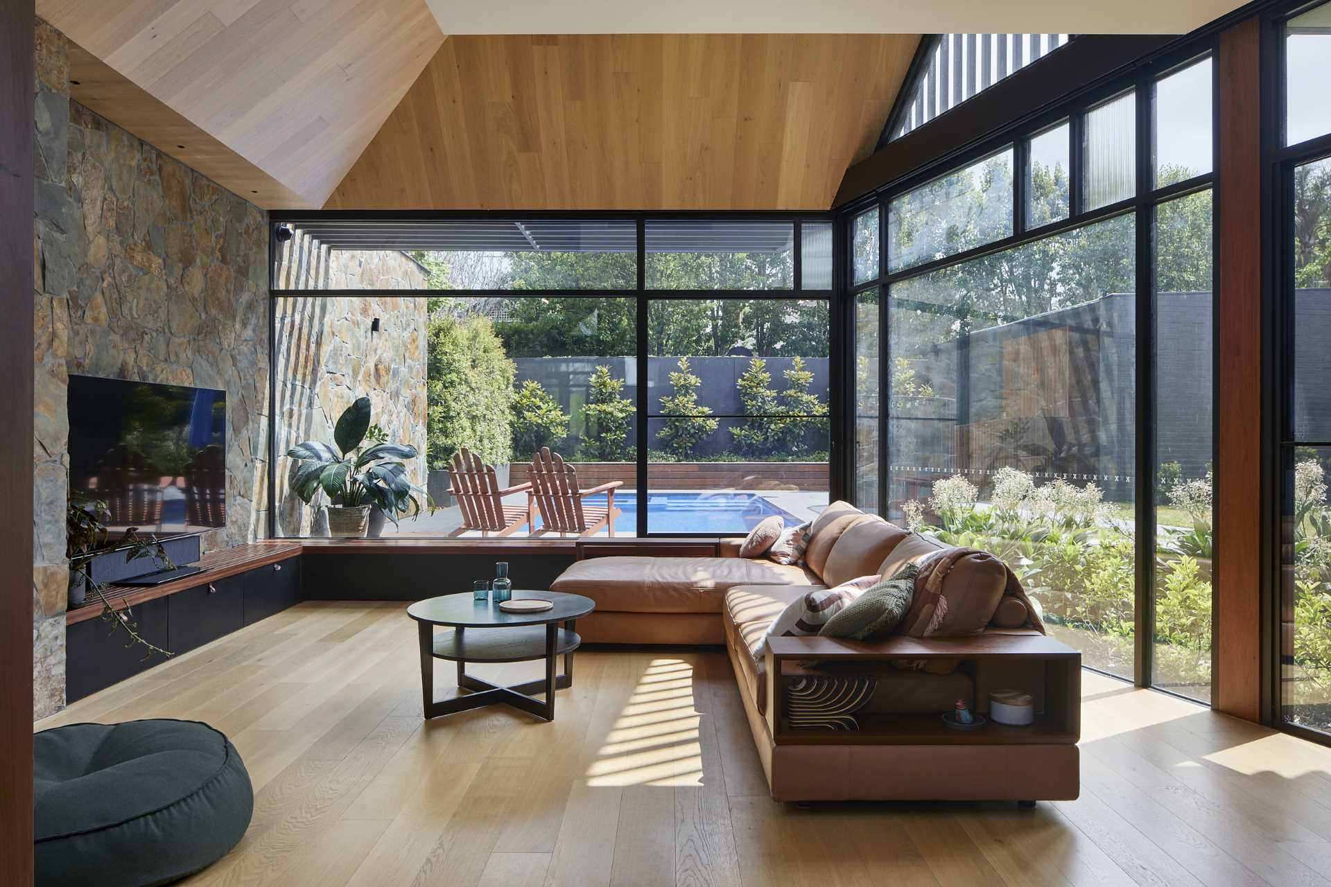 Inside this modern home, the stone wall by the swimming pool as well as the wood bench by the planter continue through to the living room which is filled with natural light from the windows.