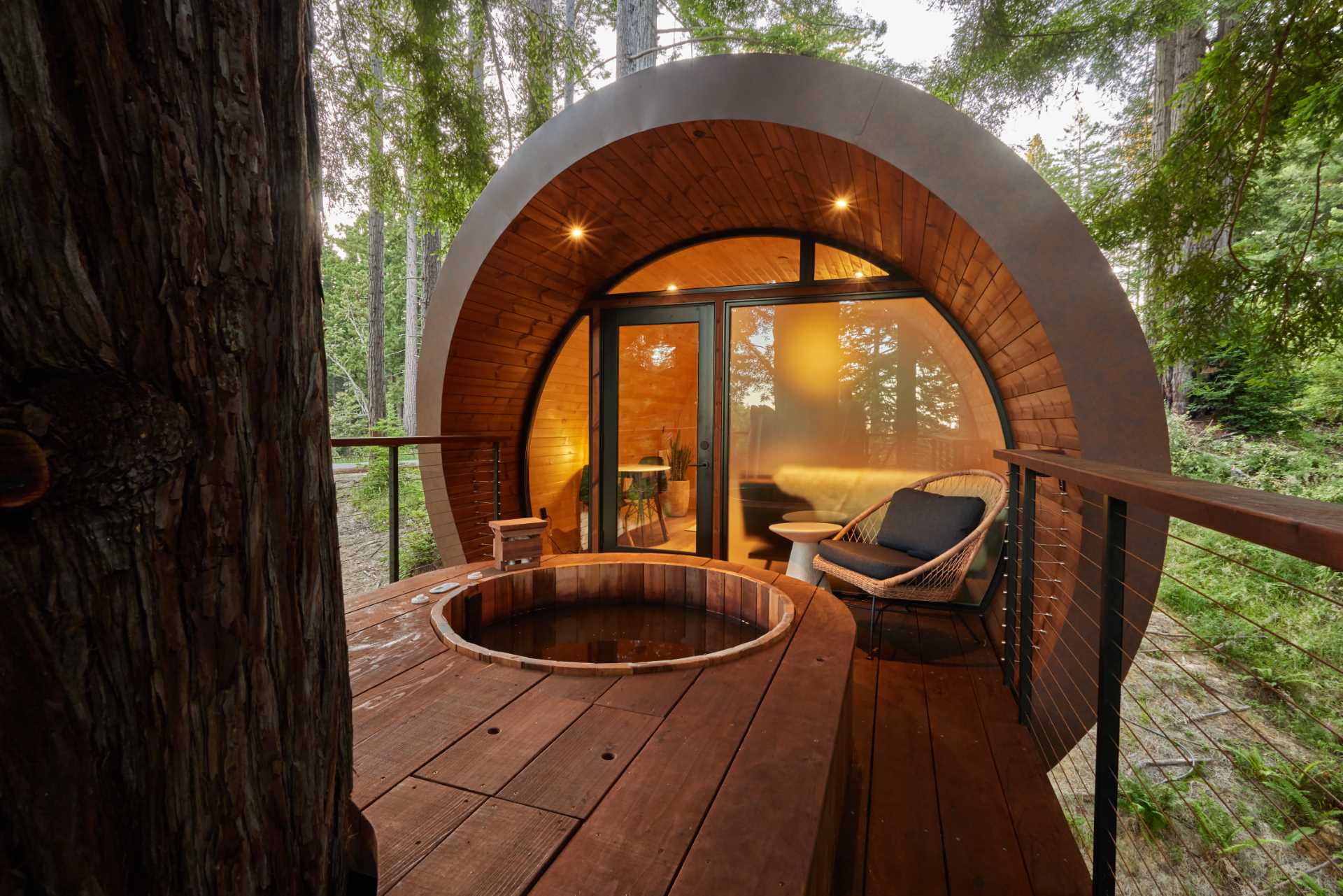 This modern treehouse cabin opens to a partially covered deck that wraps around a large tree, and has a cedar hot tub that overlooks the forest ravine.