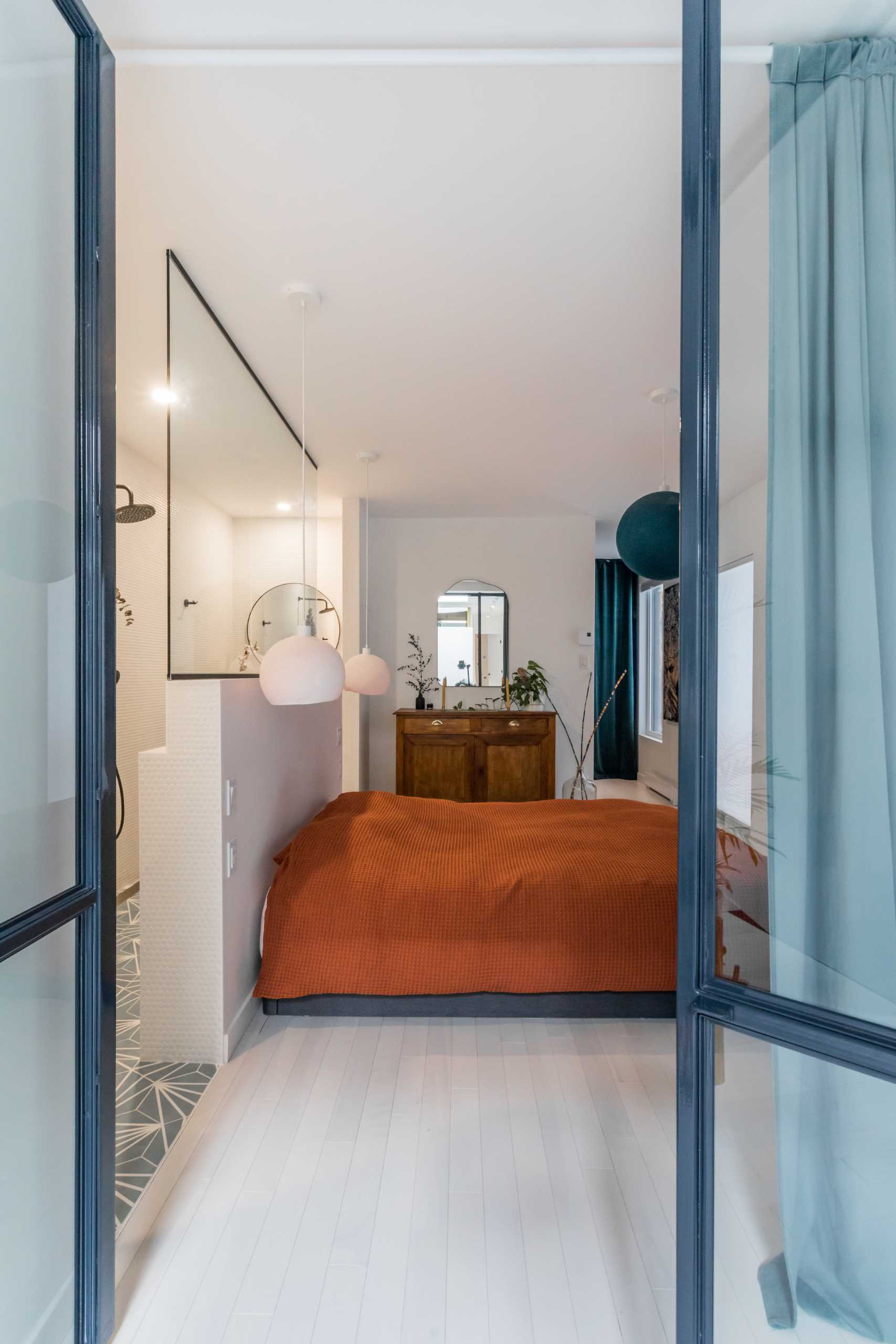Metal-framed glass doors open to reveal the primary bedroom and bathroom. A pony wall separates the sleeping area from the bathroom, where the shower separates the two vanities.