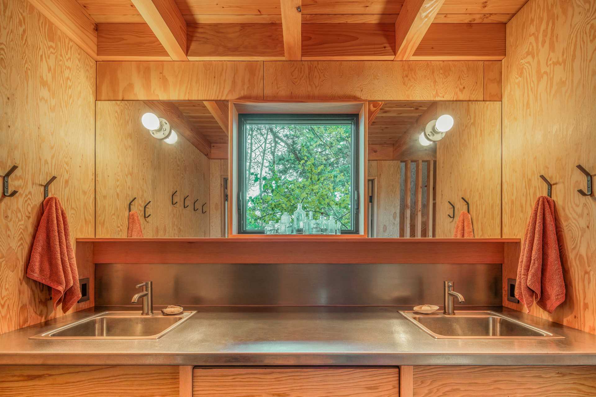 In the bathroom area of this cabin, there's a custom stainless steel counter with matching sinks, polished chrome faucets, and mirrors positioned on either side of the window.