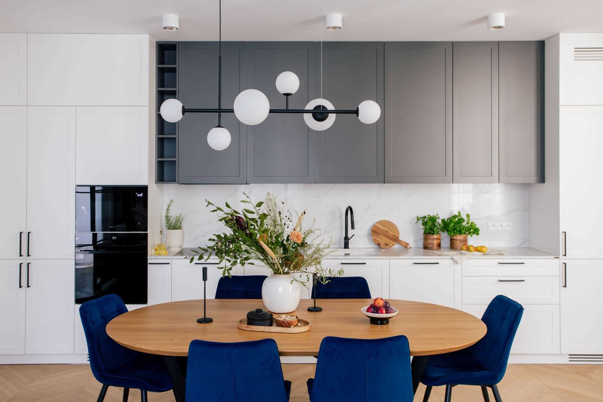 This modern dining area features an oval table with blue upholstered chairs, while the kitchen includes white and grey cabinetry.