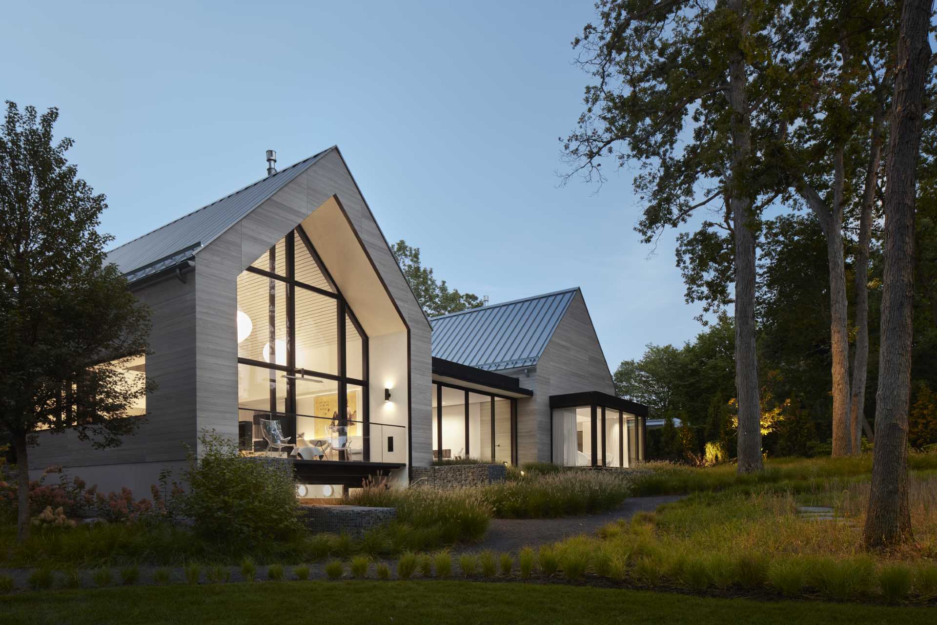 The exterior of this modern home with two gables, is composed of warm gray Accoya siding, deeply inset windows, and zinc-colored standing seam roofing. 