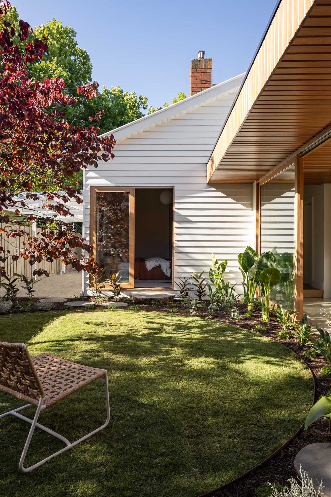 On one side of a hall with sliding glass doors, there's an outdoor sitting area with a curved grassy area, while on the other side of the walkway, there's a wood deck set up for outdoor dining.