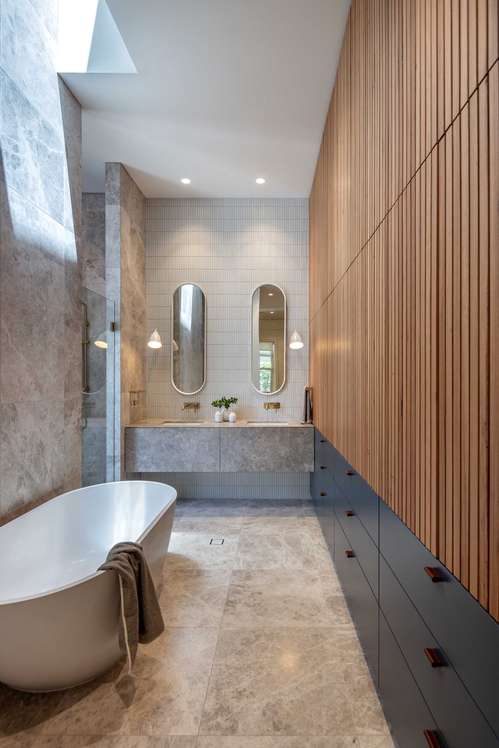 In an ensuite bathroom, tiles adorn the walls, while the bathtub is positioned underneath a skylight, a wall of wood slats adds a sense of warmth, with storage below.