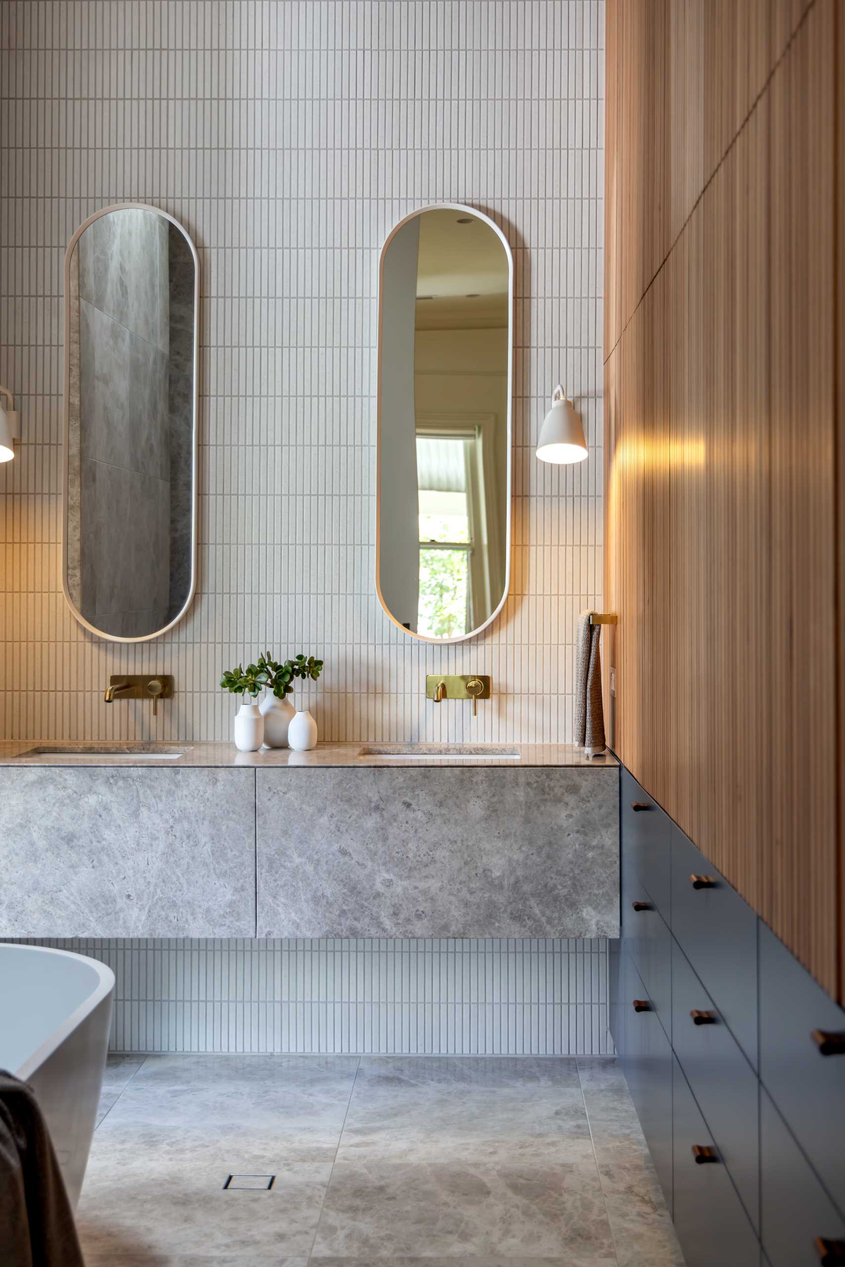 In an ensuite bathroom, tiles adorn the walls, while the bathtub is positioned underneath a skylight, a wall of wood slats adds a sense of warmth, with storage below.
