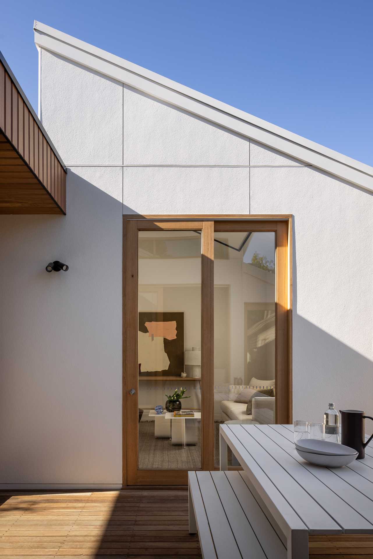 On one side of a hall with sliding glass doors, there's an outdoor sitting area with a curved grassy area, while on the other side of the walkway, there's a wood deck set up for outdoor dining.