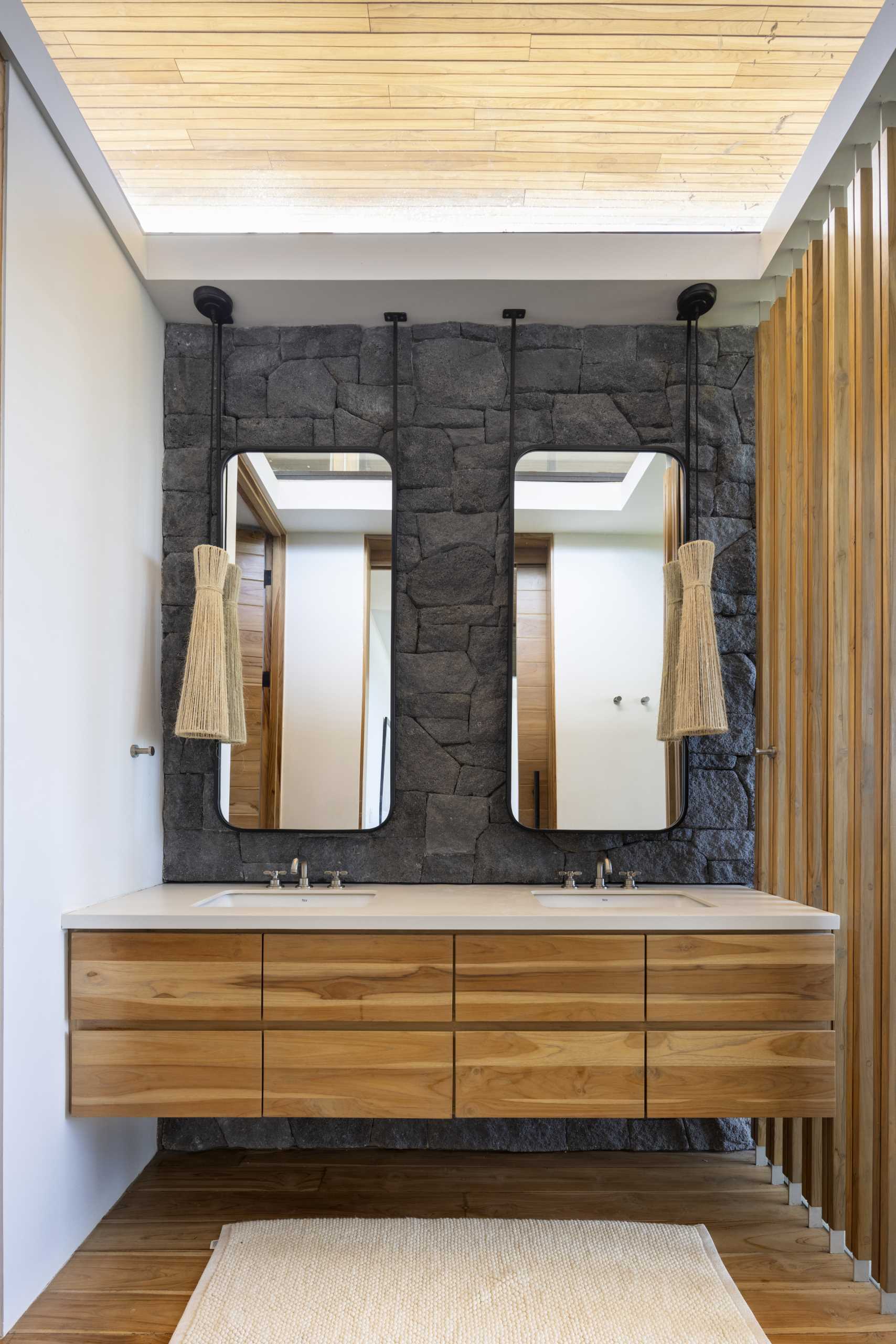 In the bathroom, a grey stone wall provides a backdrop for the wood vanity, mirrors, and pendant lights.