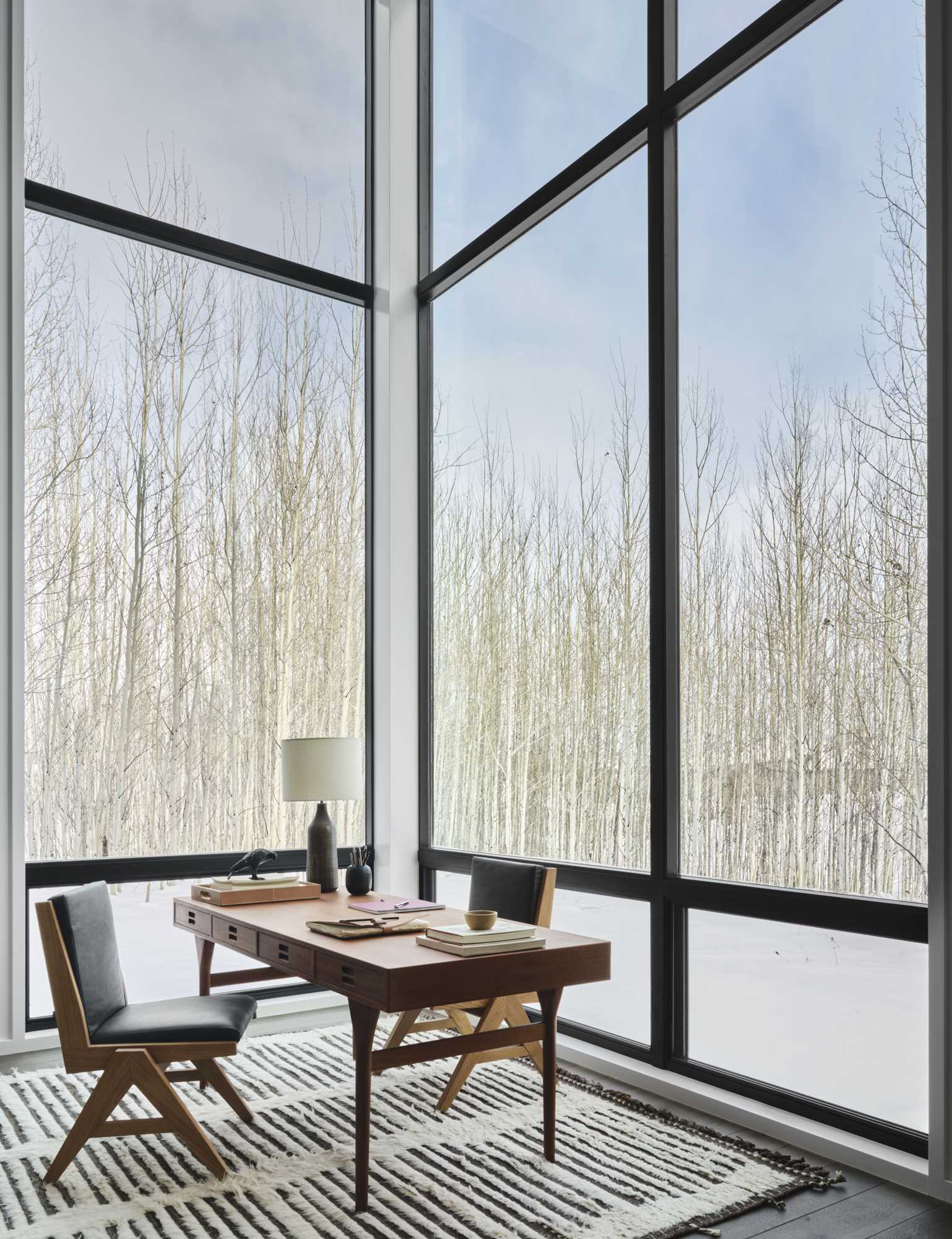 A home office is flooded with natural light from the tall black-framed windows.