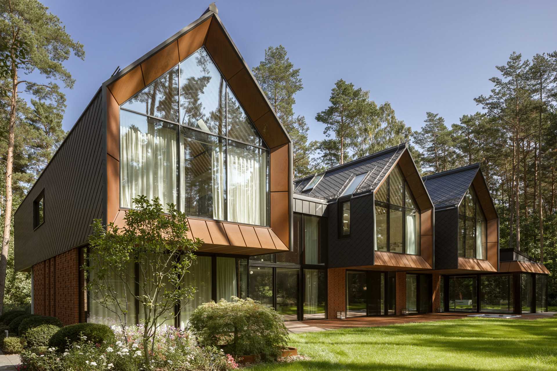 A modern home in the forest, features gabled forms, red brick, black-coated tiles, and an weathered steel on its exterior.