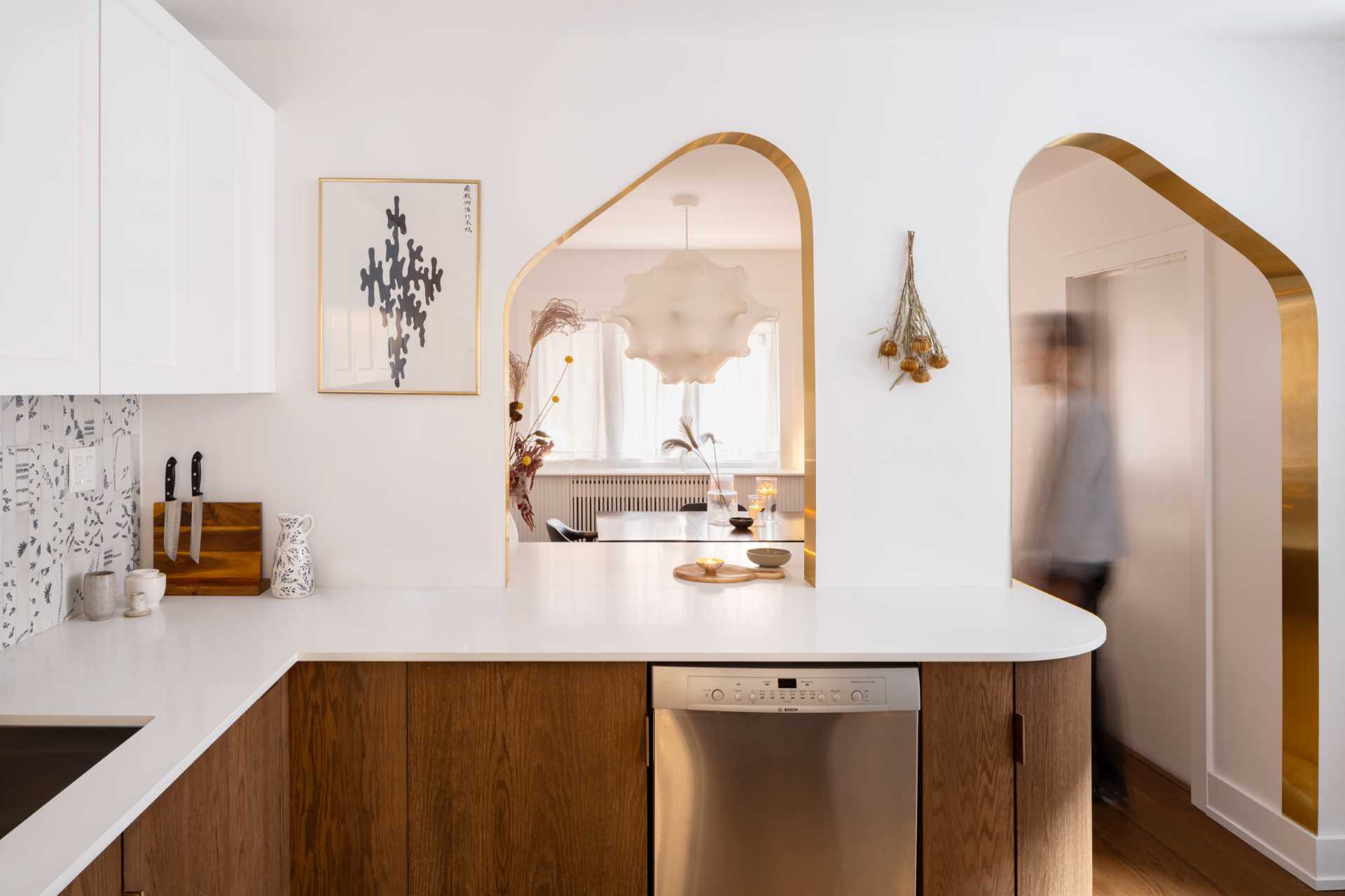 A modern kitchen with dark wood and white cabinets, white countertops, Dutch “Delft” tile, and brass-lined arches.