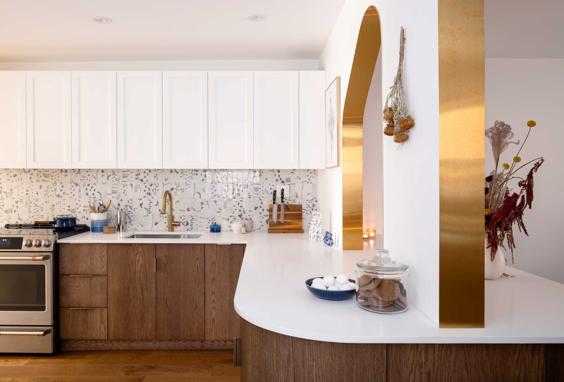 A modern kitchen with dark wood and white cabinets, white countertops, Dutch “Delft” tile, and brass-lined arches.