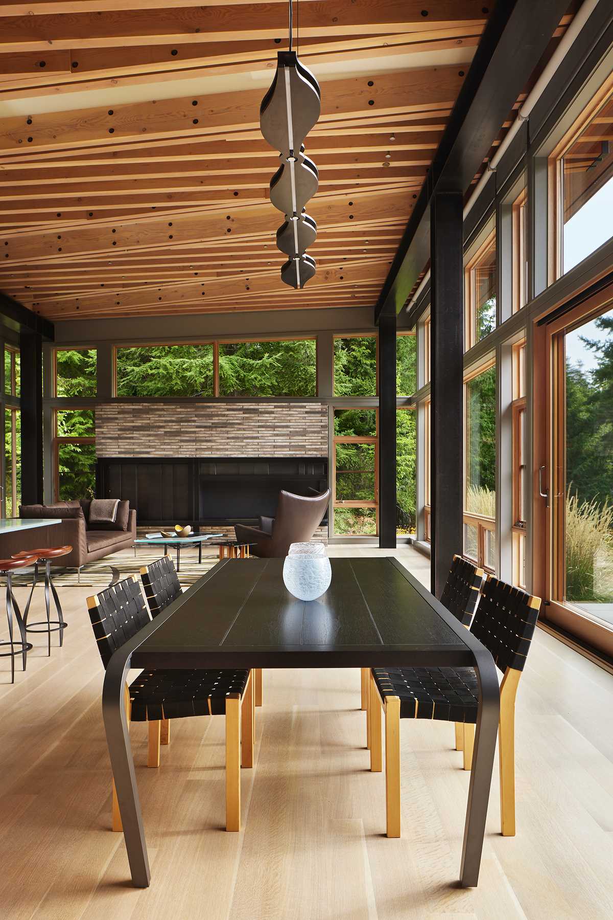 The dining table is placed across from the kitchen, with large sliding glass doors opening to a raised terrace overlooking the meadow. Custom lighting is also featured throughout the interior, like above the dining table, and in the kitchen.