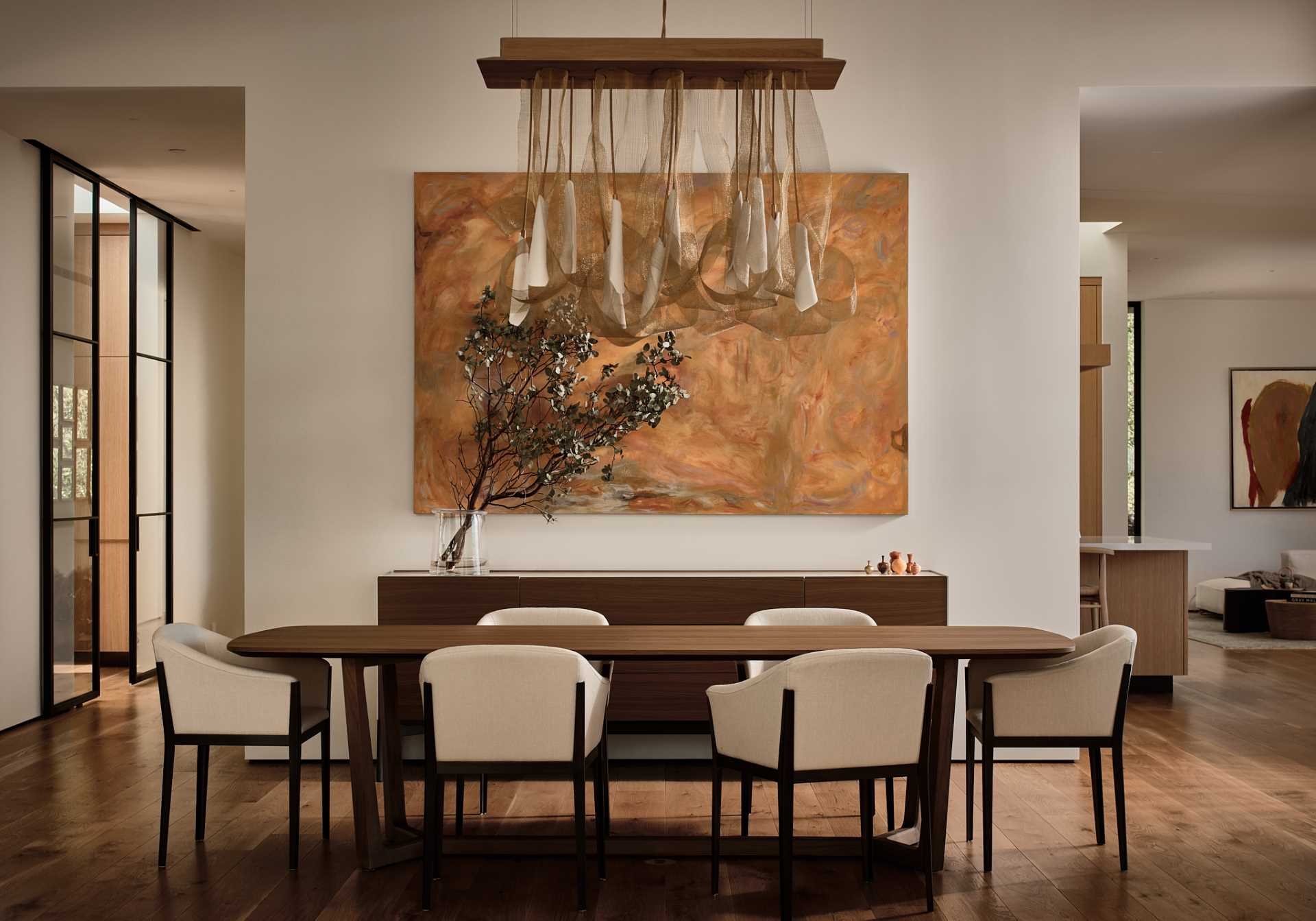 In a formal dining area, a wood table is surrounded by contemporary chairs, while a sculptural light hangs above the table and a large art piece hangs on the wall.